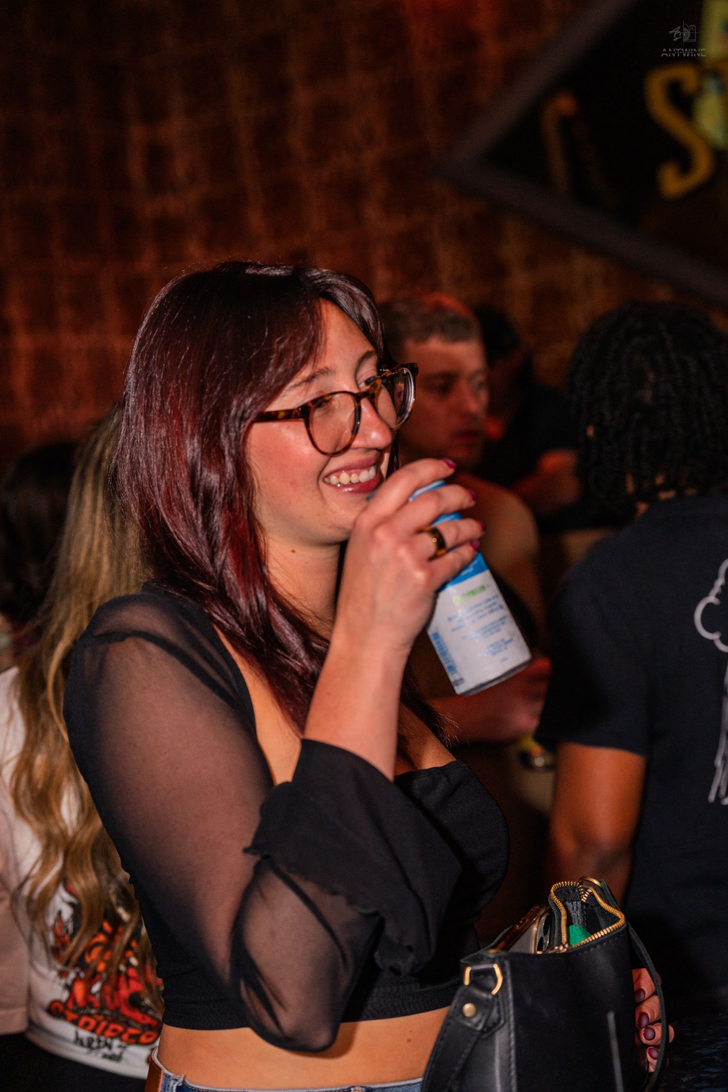 A smiling young woman with glasses drinks from a can at a lively nightclub event.