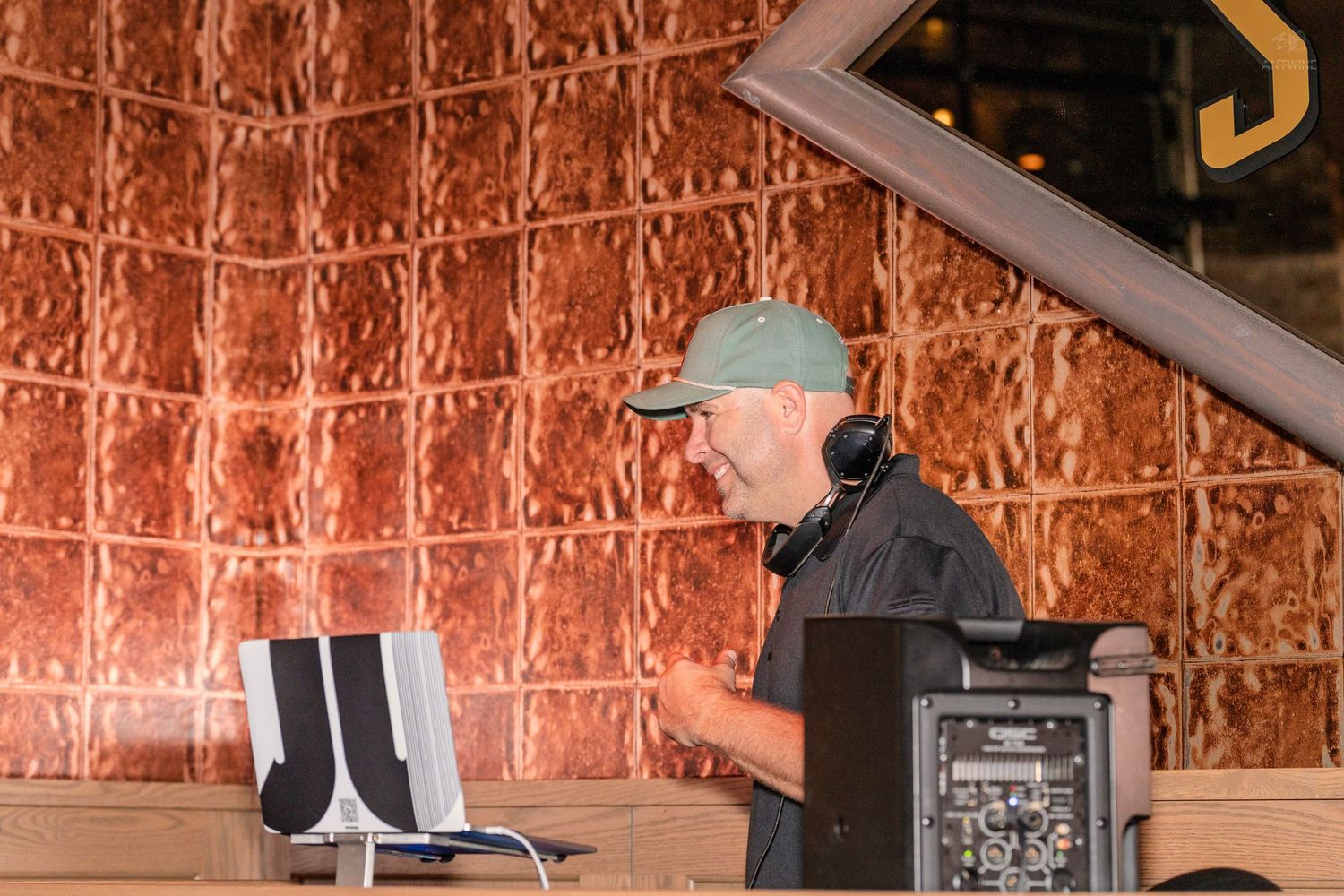 Male DJ wearing headphones and a cap performing at an indoor venue with copper wall decor.