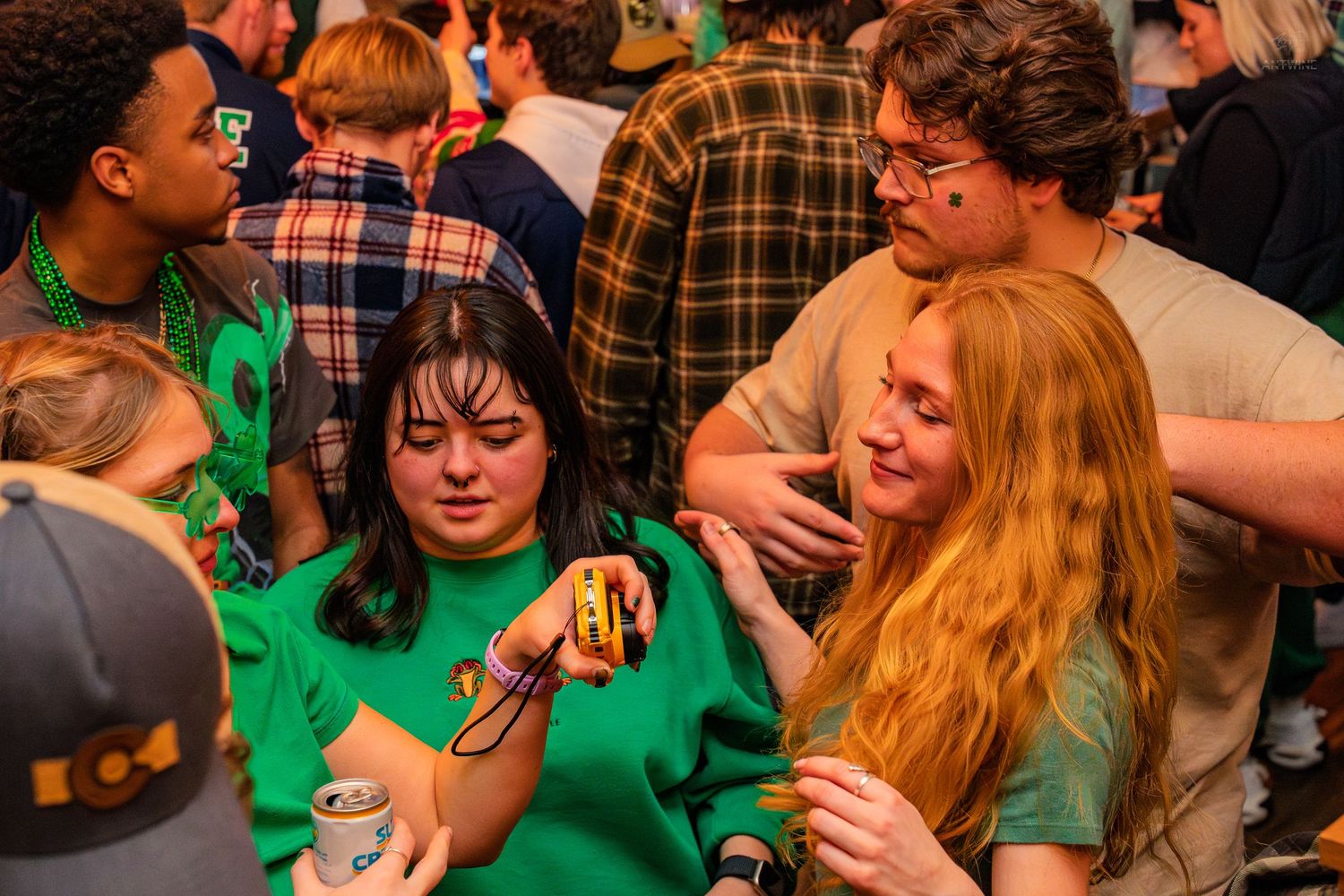 Two women in green shirts examining an item at a crowded St. Patrick's Day bar celebration.