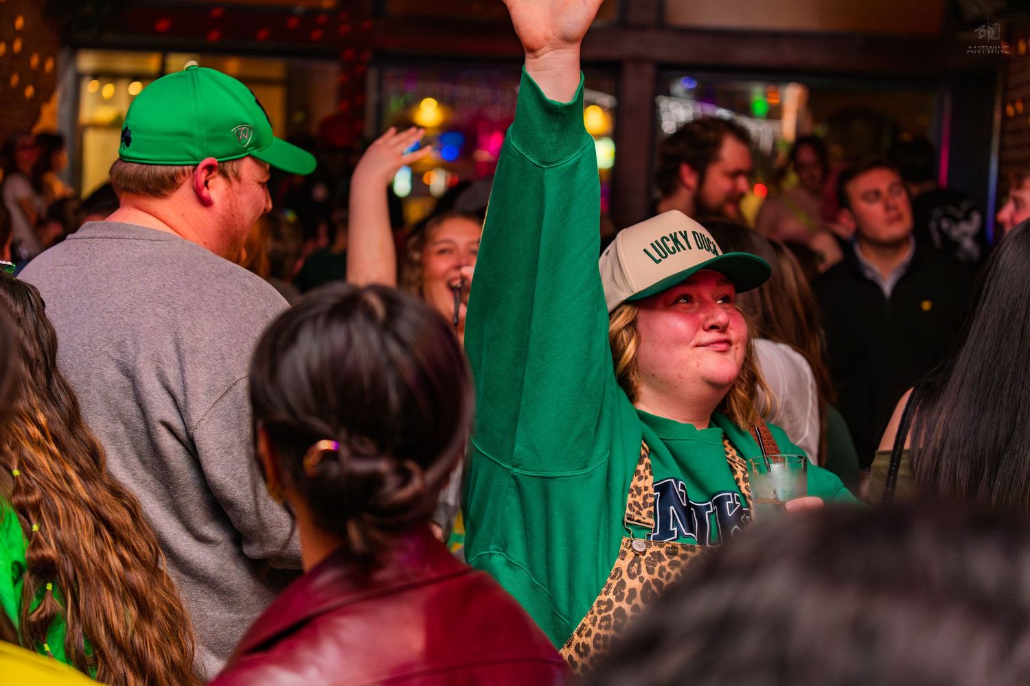 Excited woman in green Lucky hat dances with arm raised at St. Patrick's Day party.