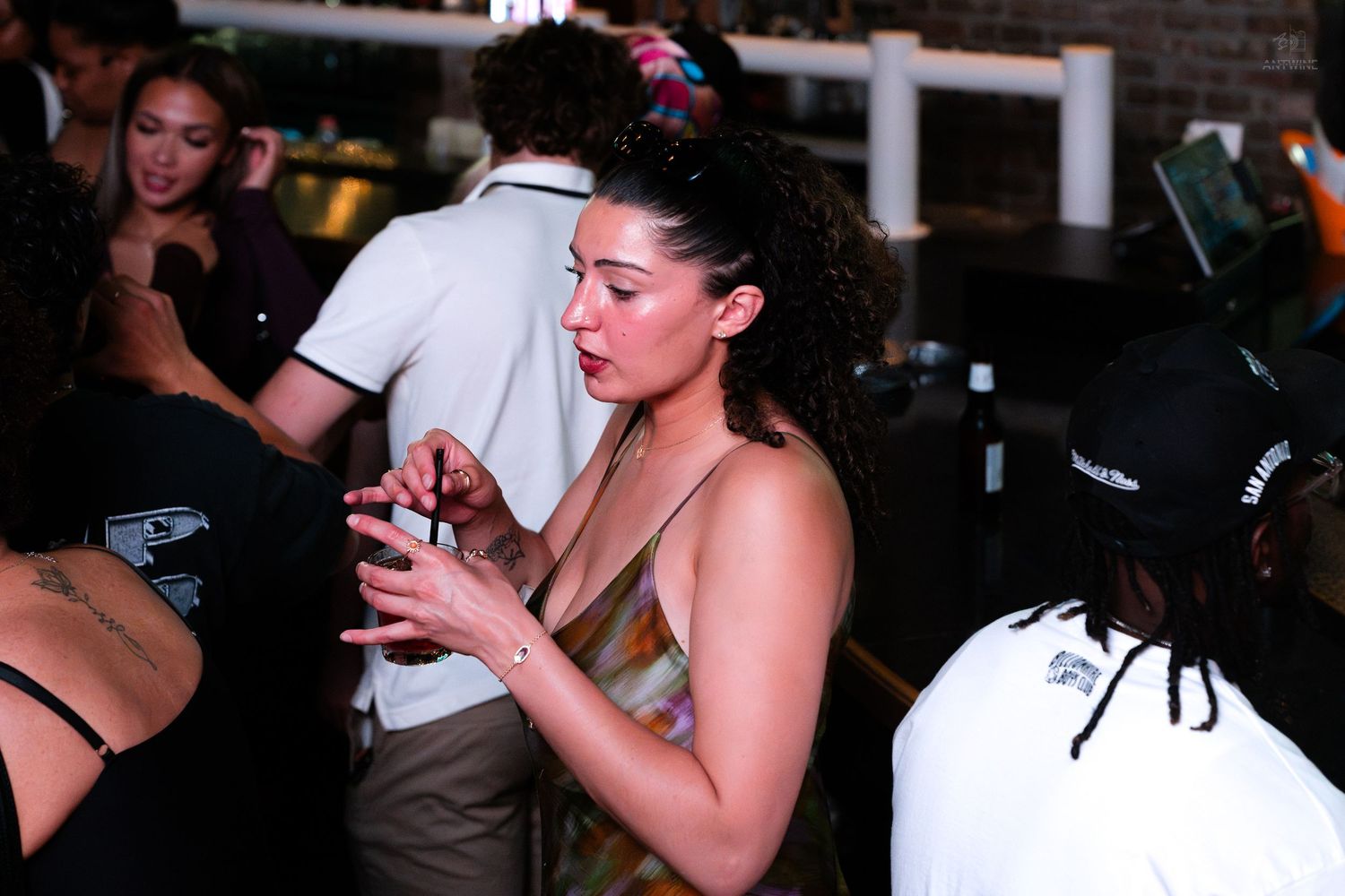 Young woman in a gold dress checking her phone at a busy bar or nightclub.