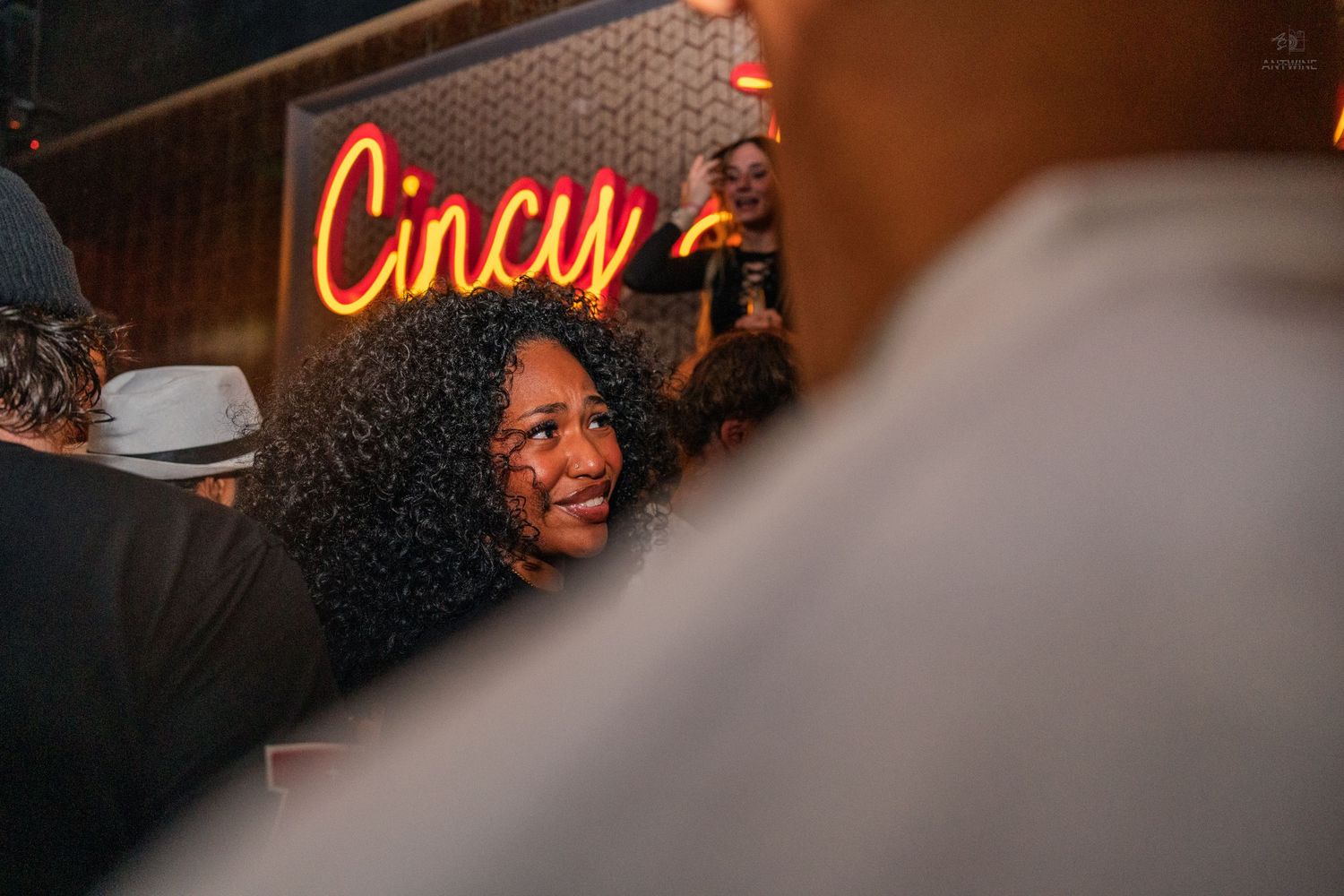 A woman with curly hair smiles at a Cincinnati Love neon sign bar event.