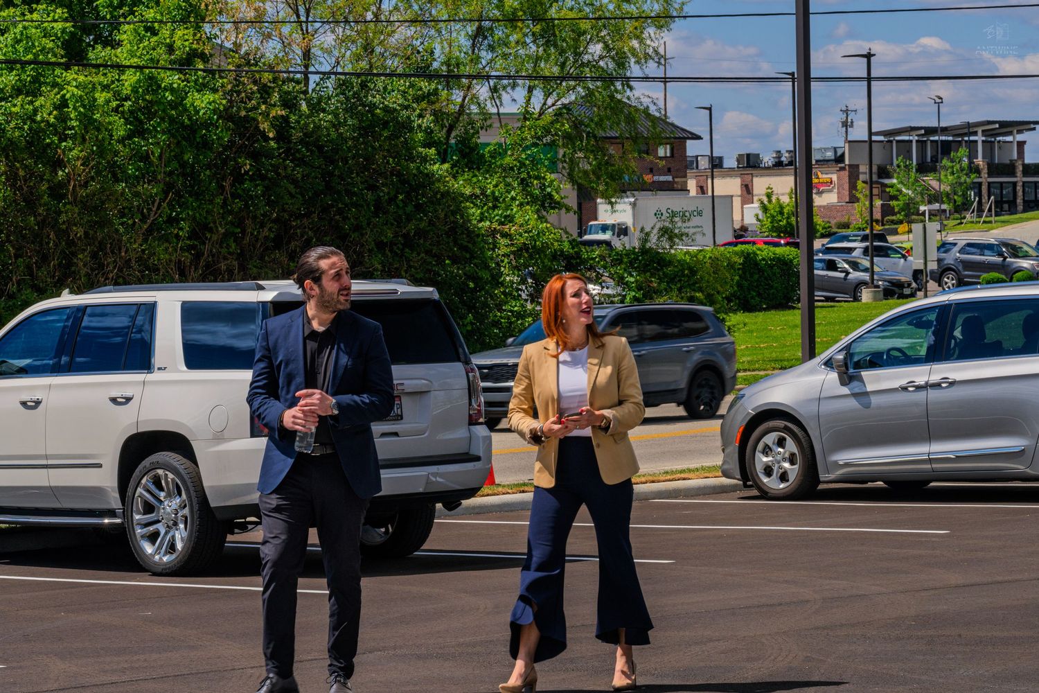 Two professionals walking through a sunny parking lot, man in navy suit and woman in tan blazer with blue pants.