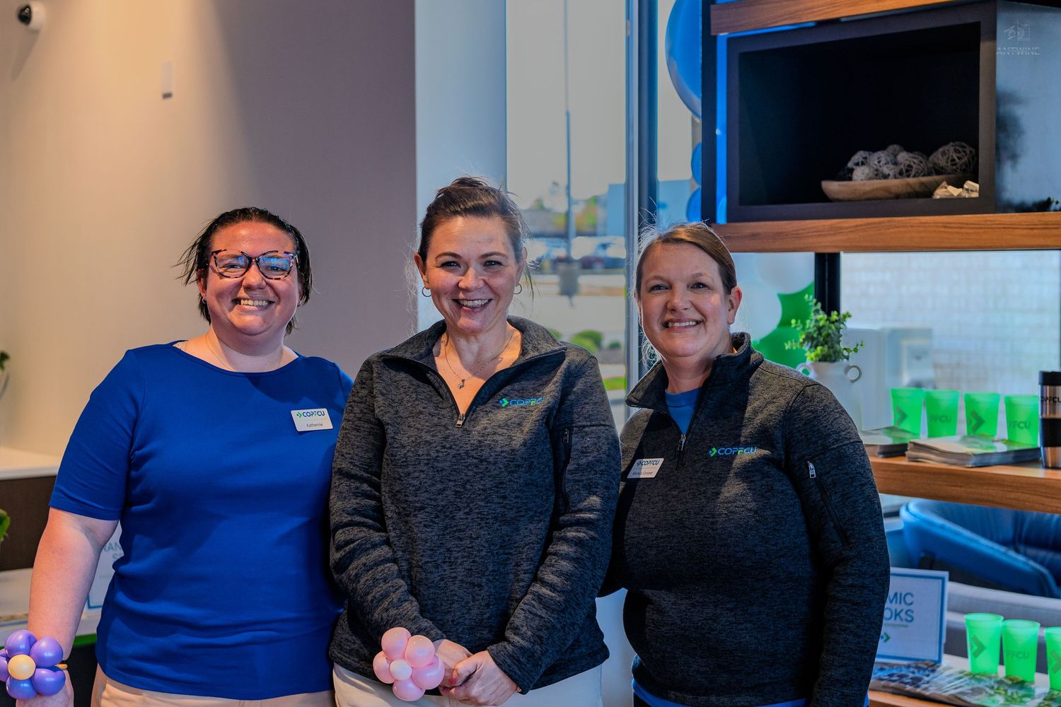 Three smiling women in blue tops pose together inside a modern retail or office space with shelving.