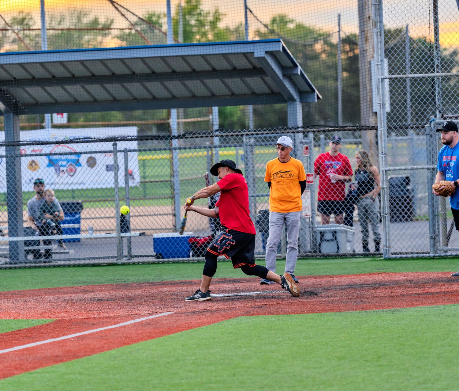 A batter in red swings at a softball pitch during an evening recreational league game on a well-maintained diamond.
