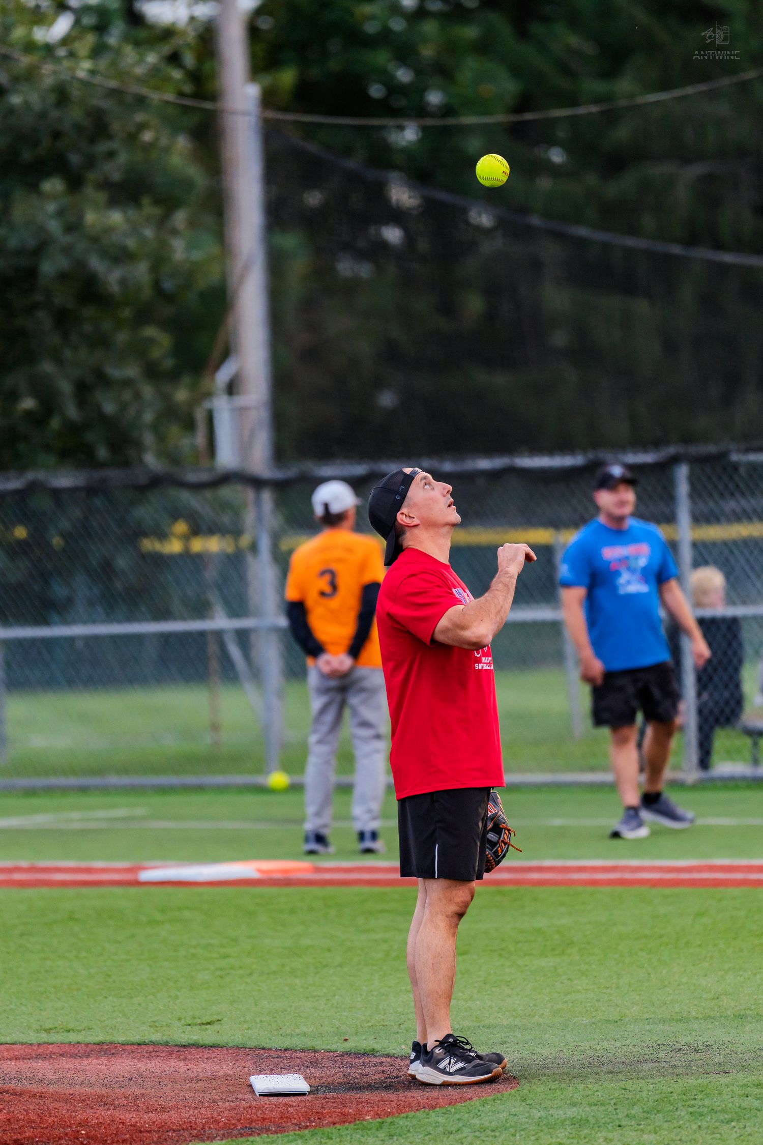 Softball player in red shirt tossing a yellow ball upward near the pitcher's mound during a recreational game.