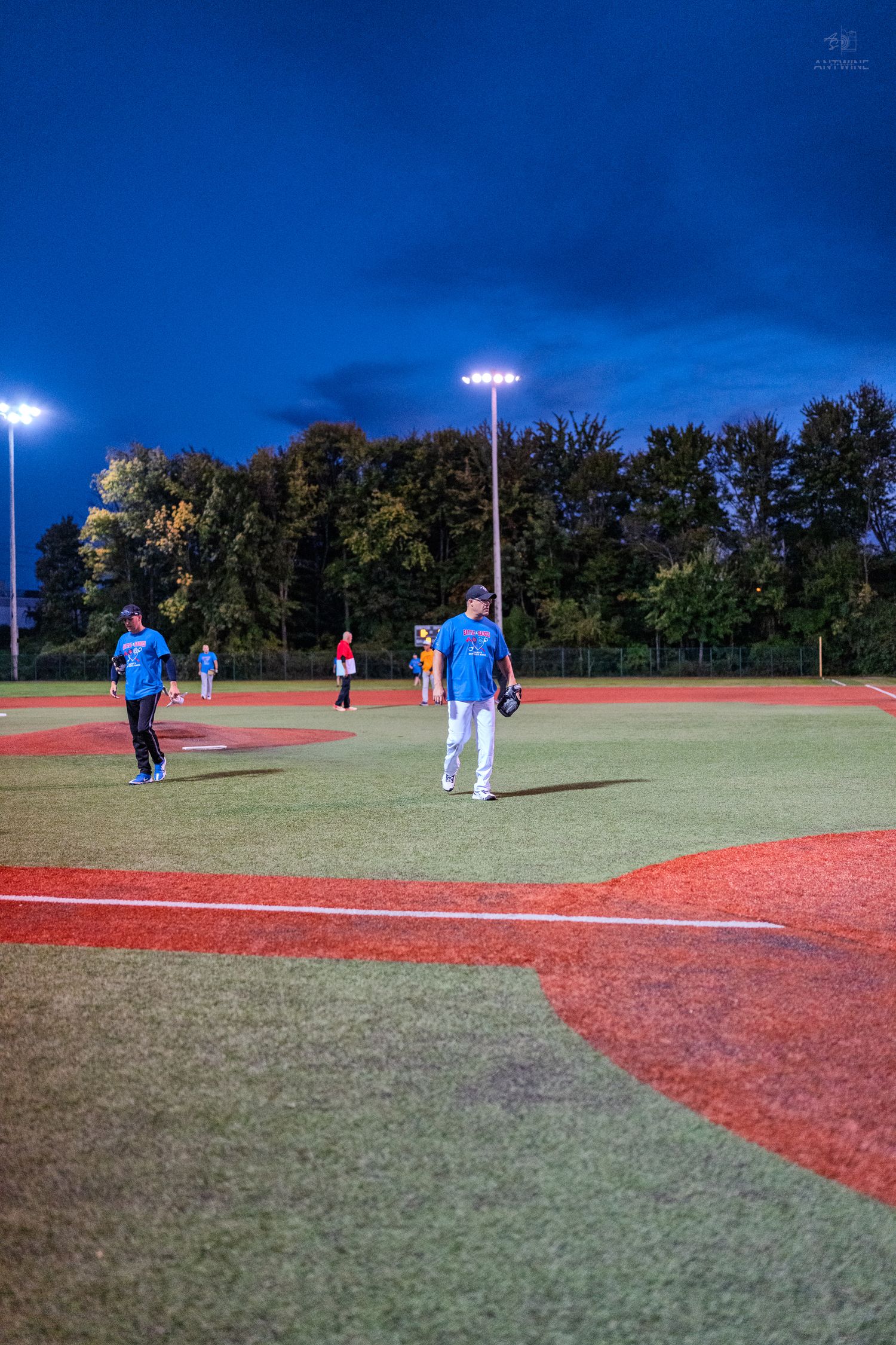 Baseball players in blue uniforms on a lit field at dusk with trees and stadium lights in background.