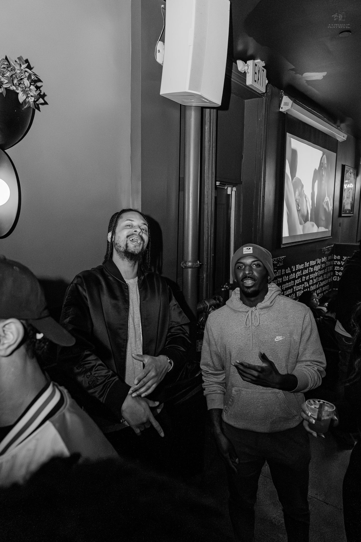 Black and white photo of two men laughing and conversing at an indoor social gathering or event.