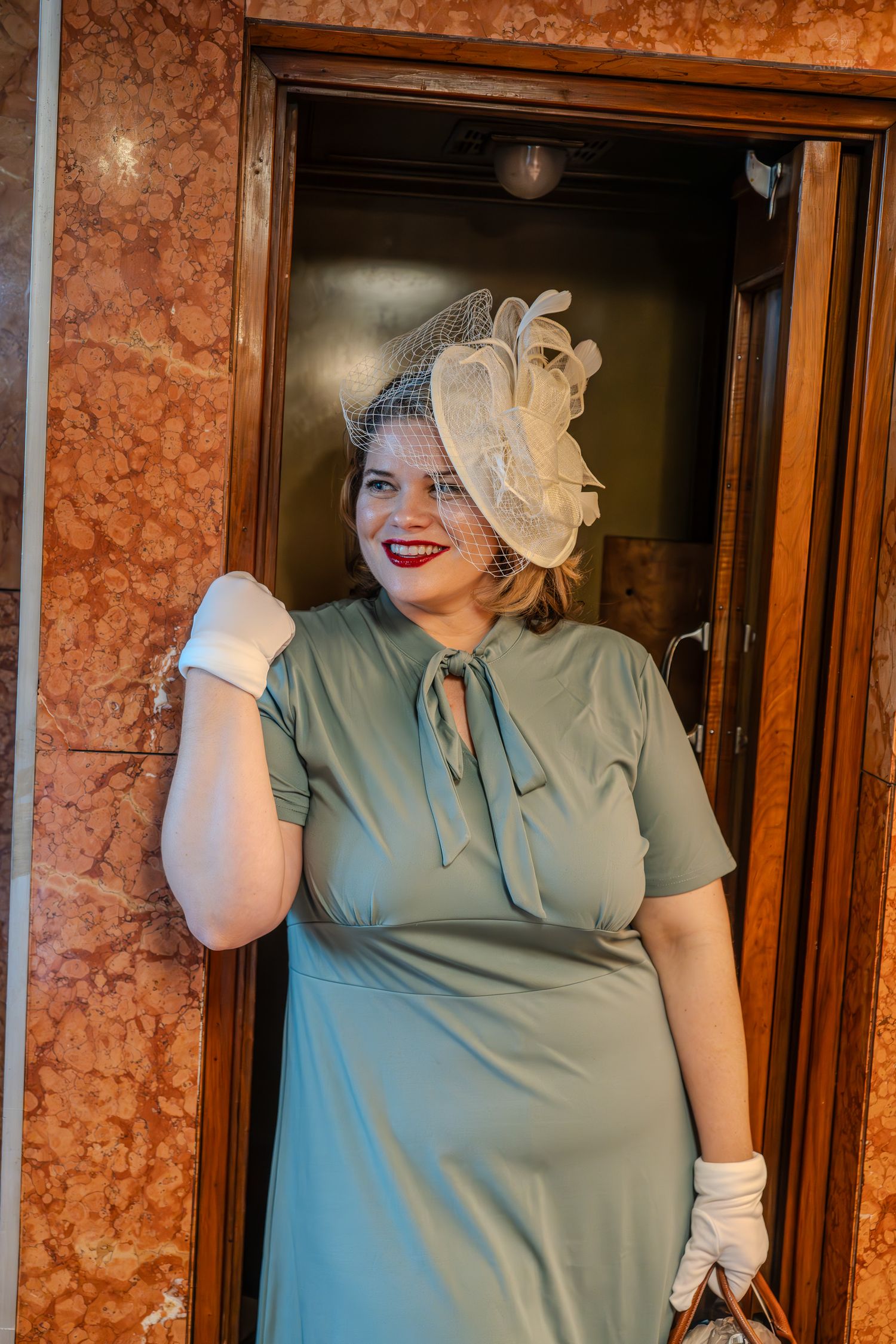 Woman in vintage sage green dress, white gloves, and fascinator hat poses elegantly by ornate marble elevator doors.