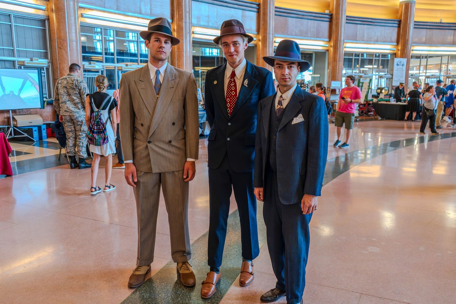Three men pose in vintage 1940s-style suits and fedora hats in a grand lobby.