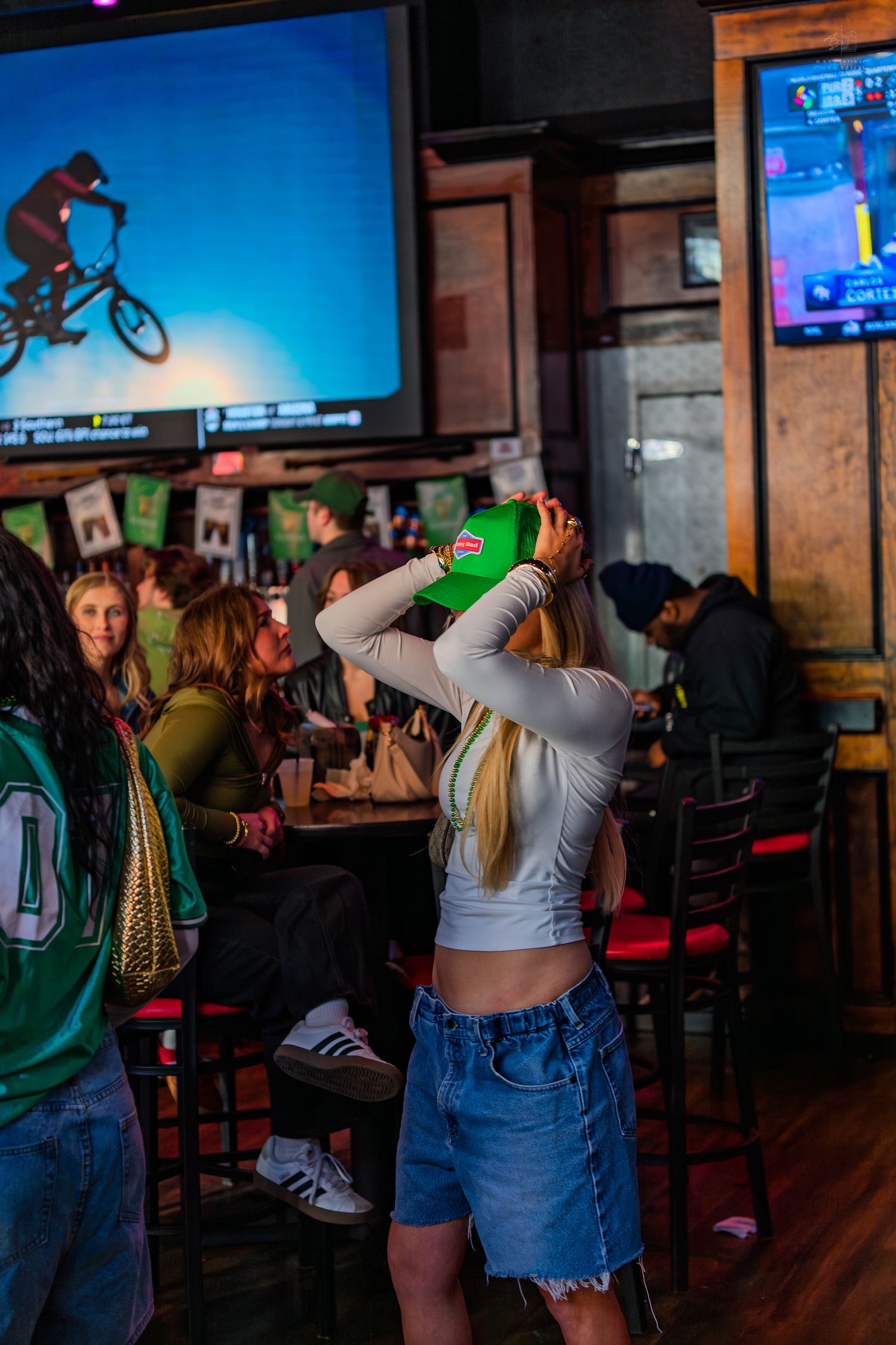A blonde woman puts on a green cap in a busy sports bar while patrons socialize and watch TV screens.