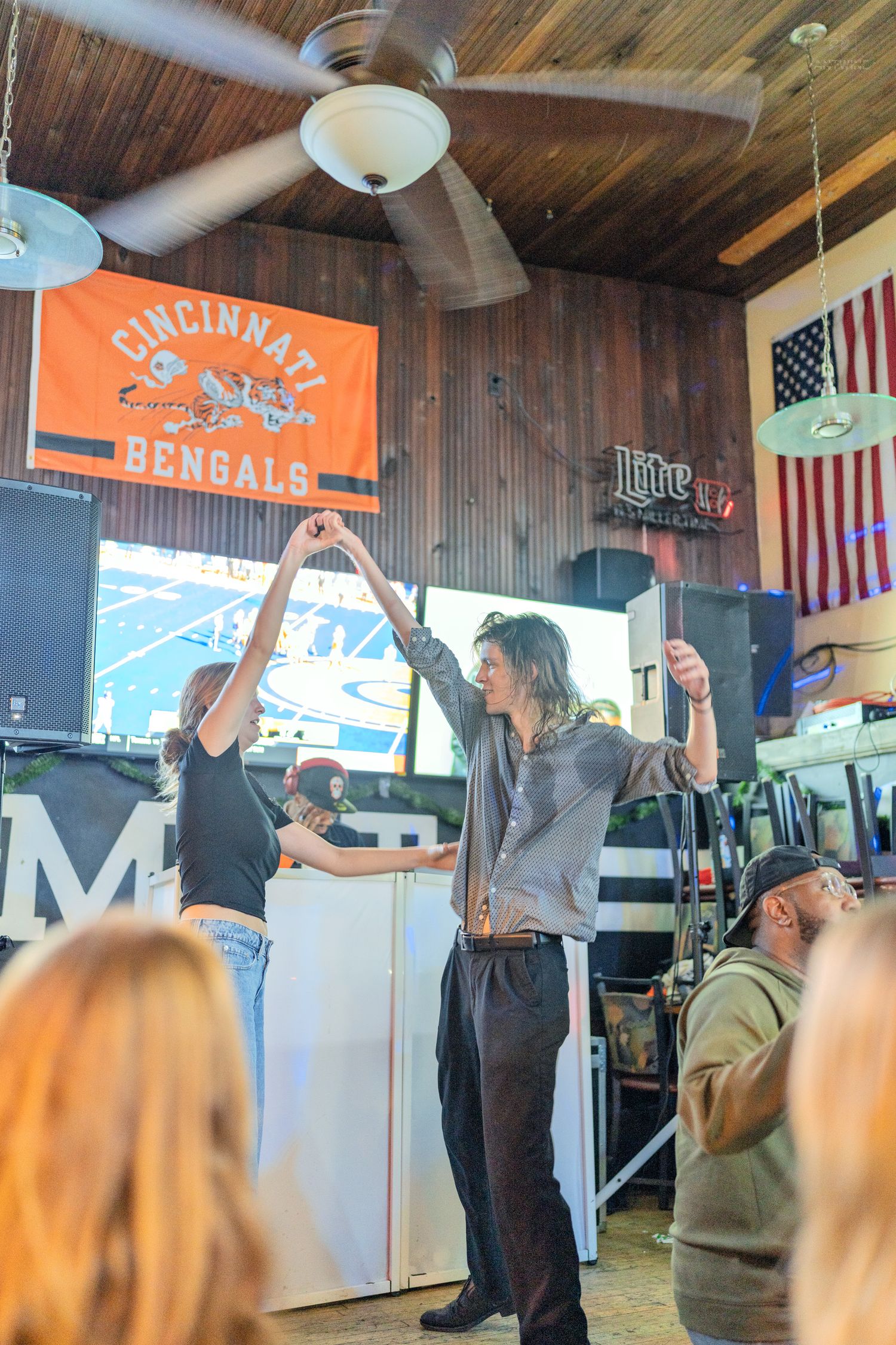 Two people dancing in a bar with Cincinnati Bengals flag, American flag, and Miller Lite sign on wood-paneled walls.