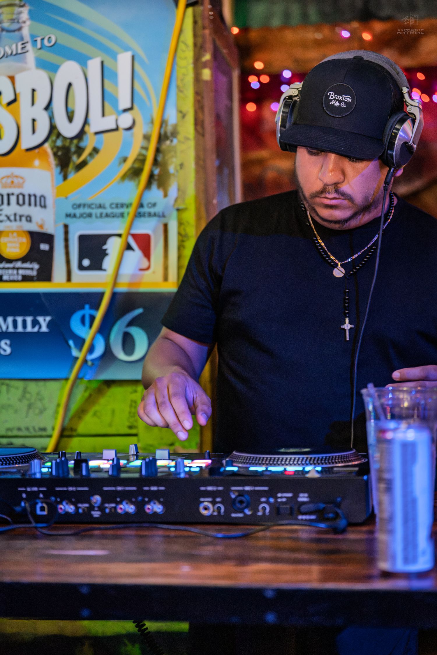 A DJ wearing headphones and a black cap operates turntables at a bar with a Corona baseball advertisement behind him.