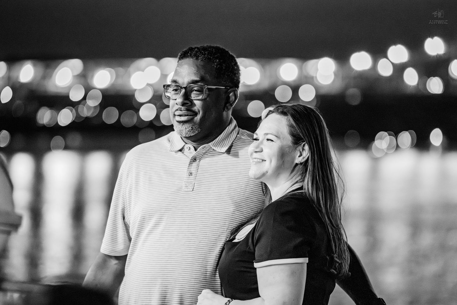 Black and white photo of a couple smiling together at night with bokeh city lights reflecting on water behind them.