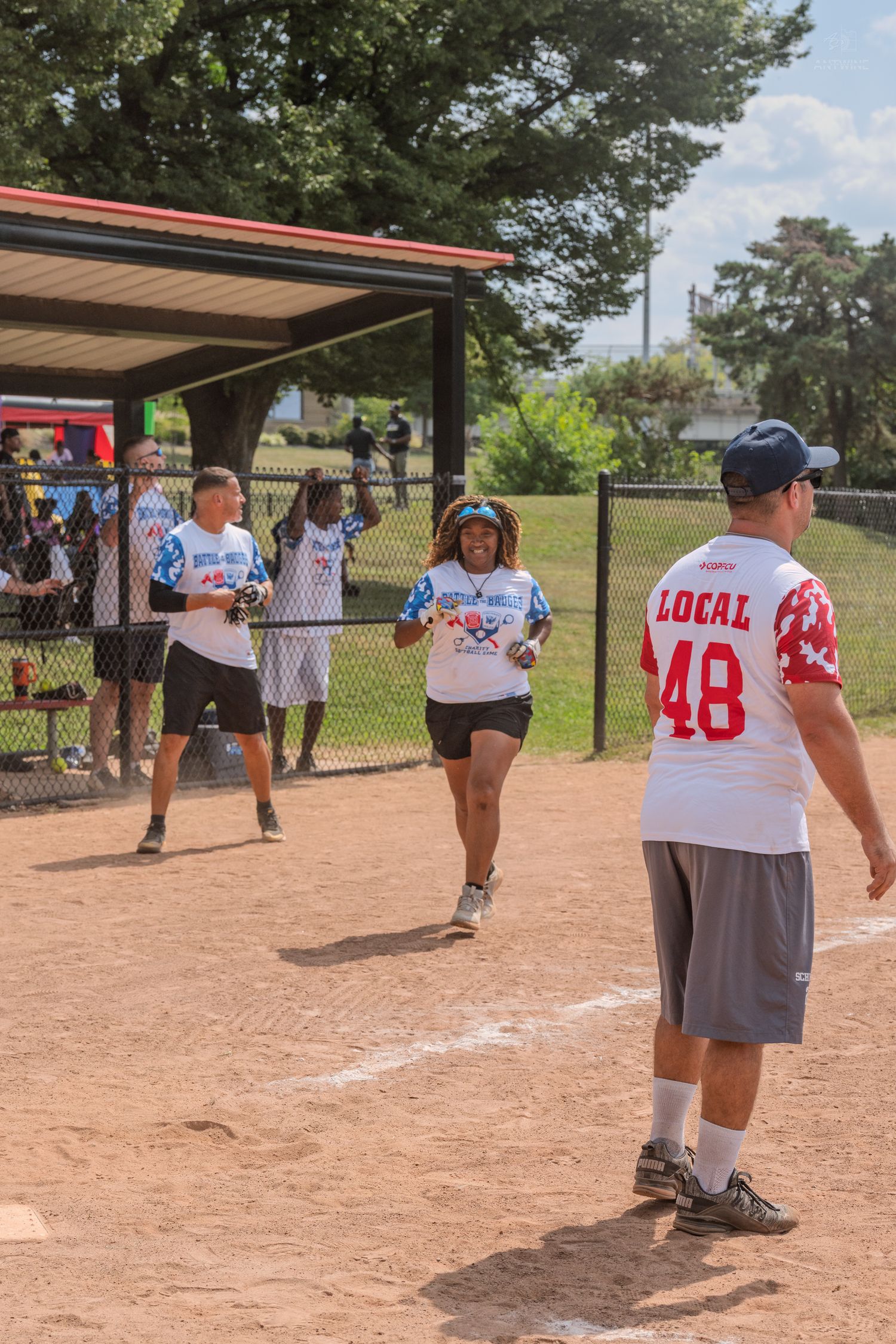 Softball players celebrate near dugout as runner approaches home plate during charity game.