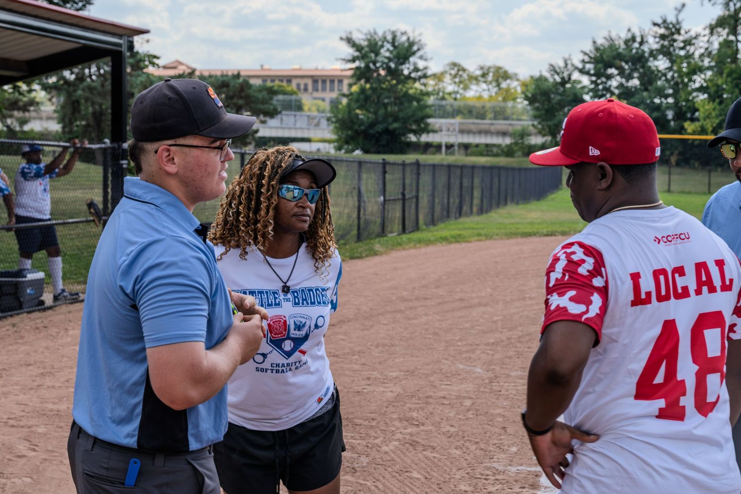 Three people in casual sports attire having a conversation near a softball field dugout.