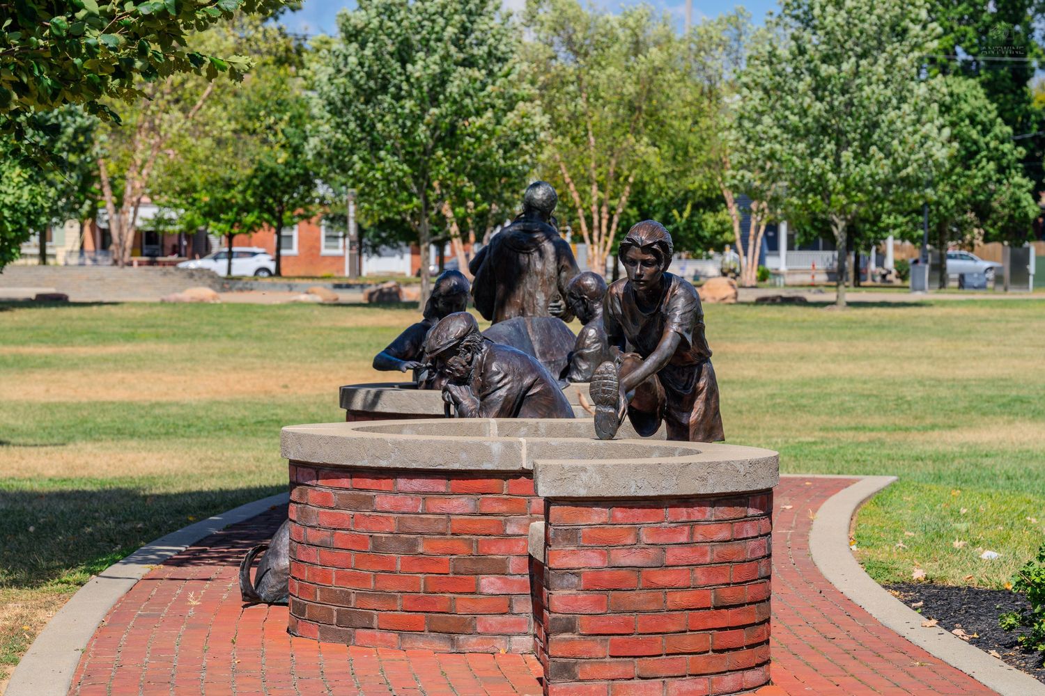 Bronze sculptures of historical figures on a brick pedestal in a sunny green park with trees in the background.