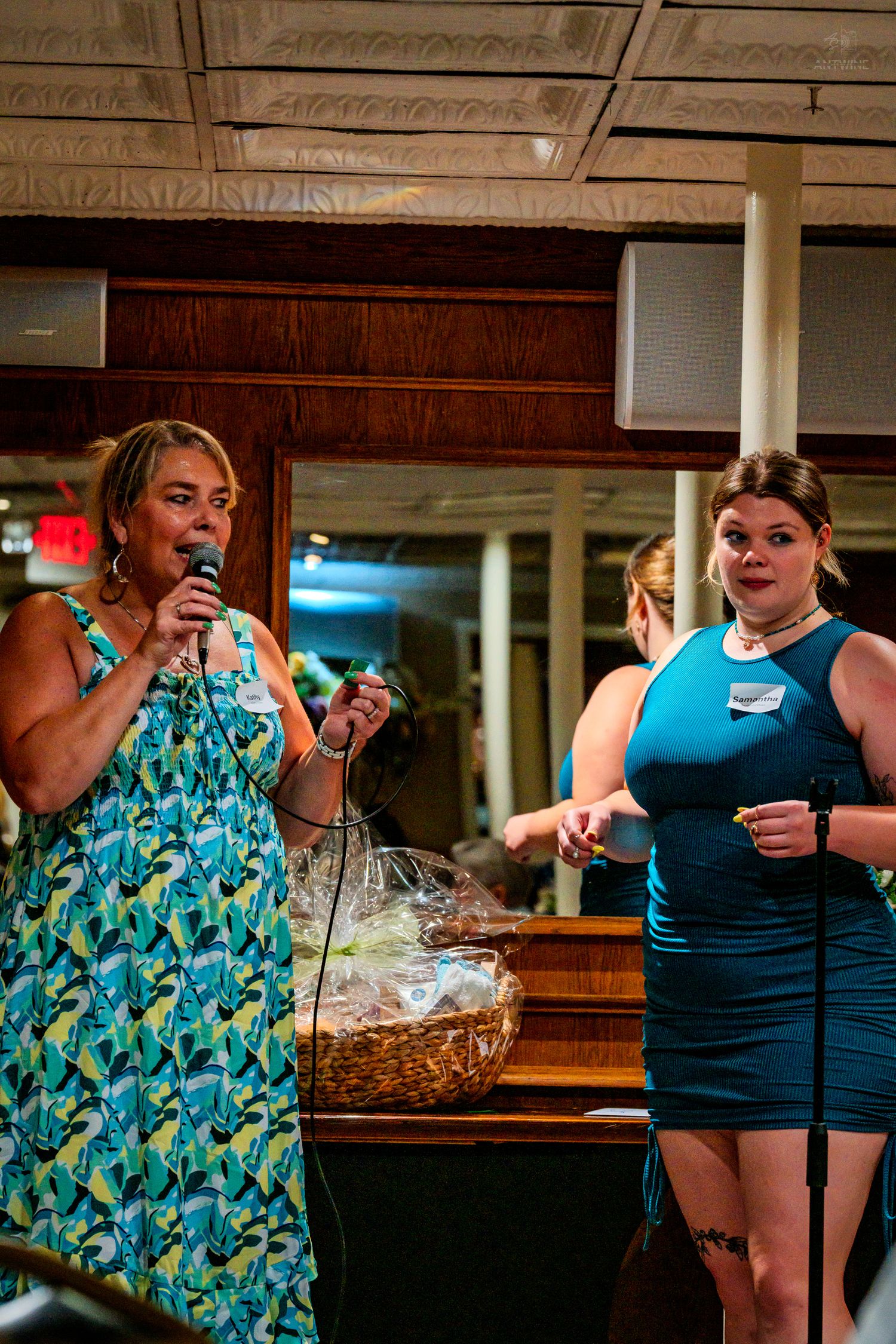 Two women in teal outfits speak at a microphone during an indoor event with a gift basket nearby.