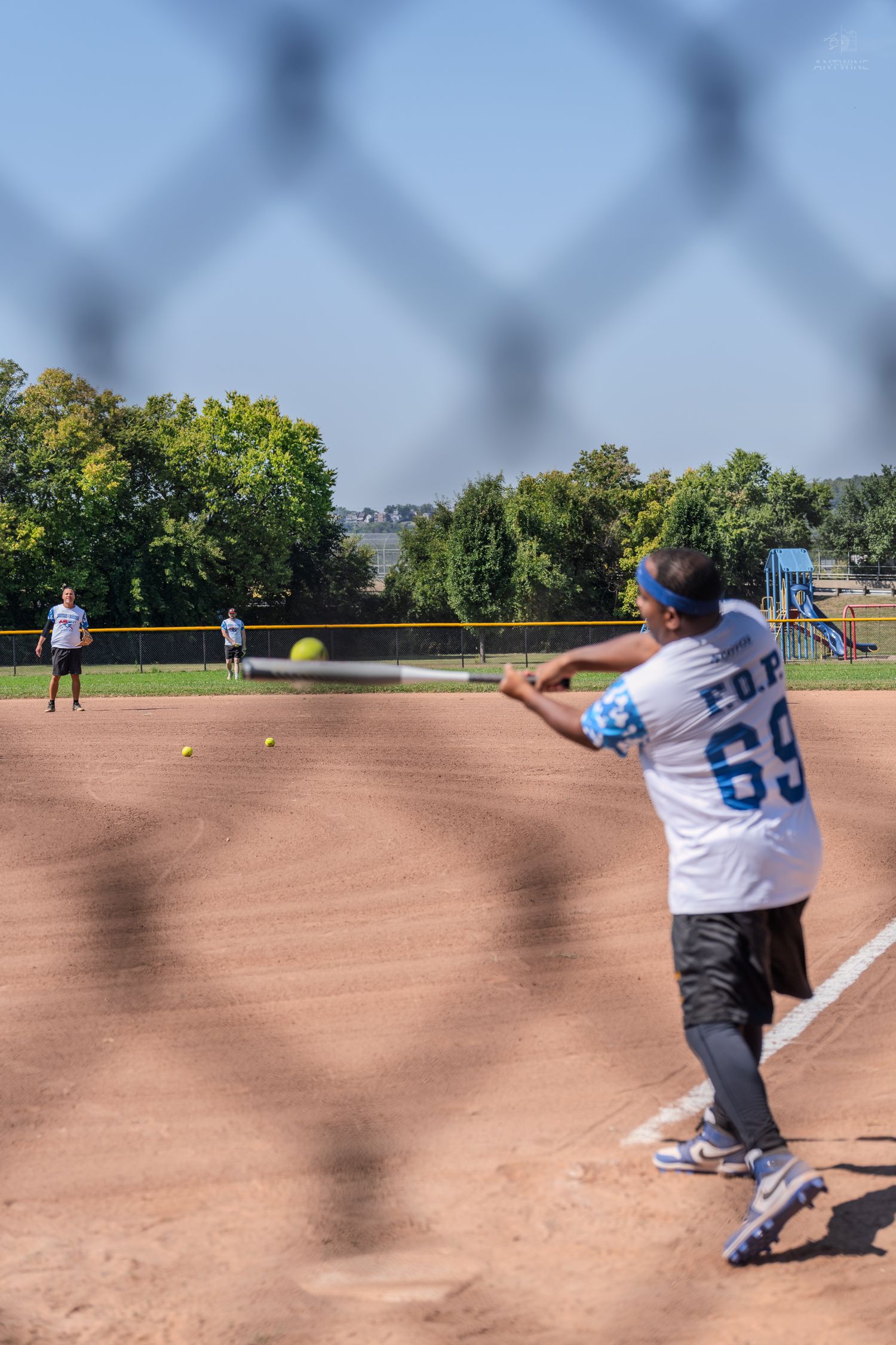 Player in jersey number 69 swings at a softball during an outdoor game on a dirt field, viewed through a chain-link fence.