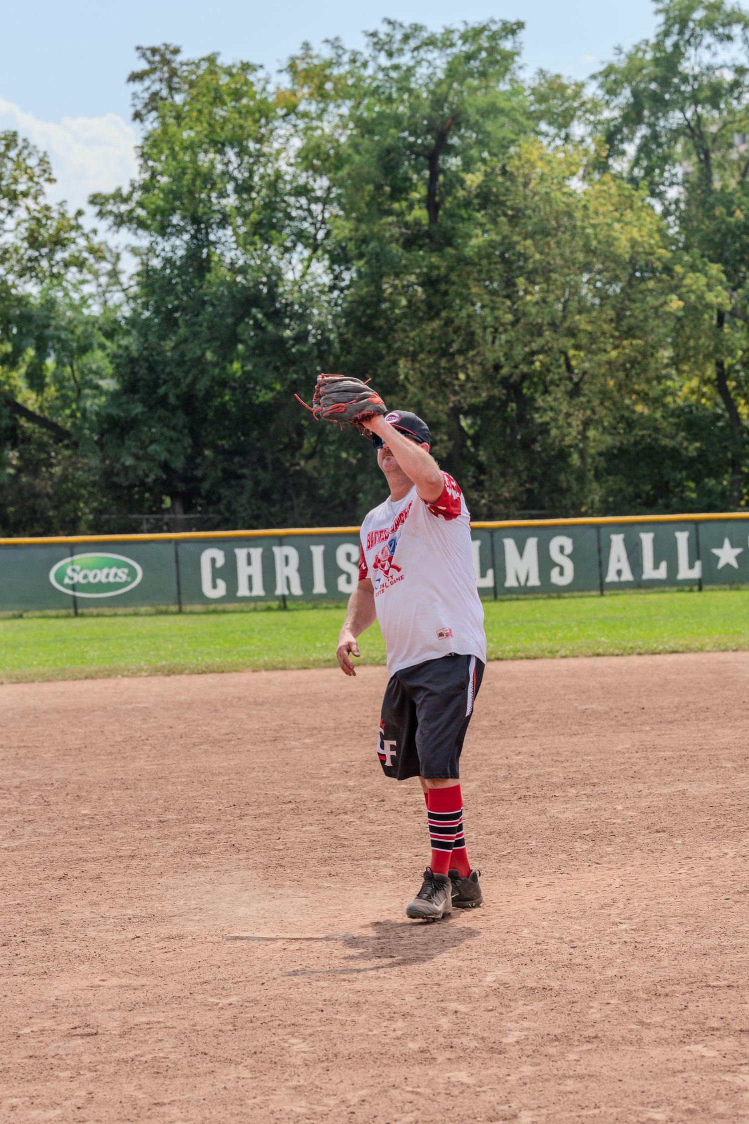 A softball player in a white jersey and red socks throws the ball on a sunny outdoor baseball field.