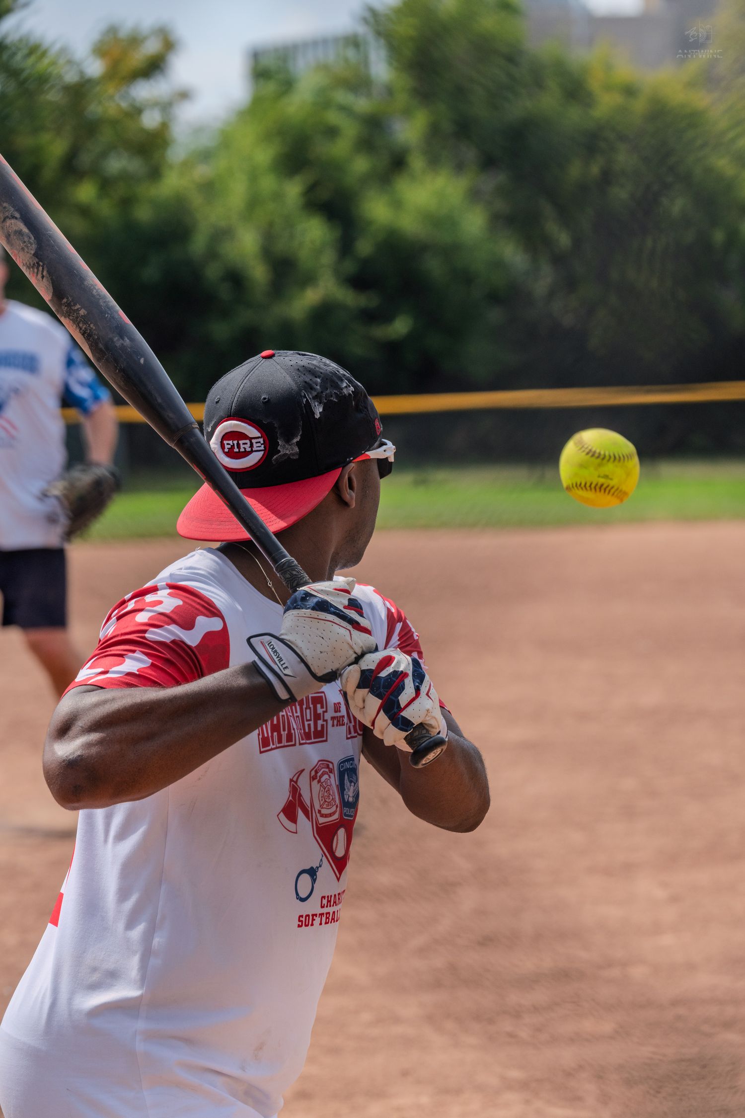 Male batter in red cap and gloves prepares to hit yellow softball during outdoor game.