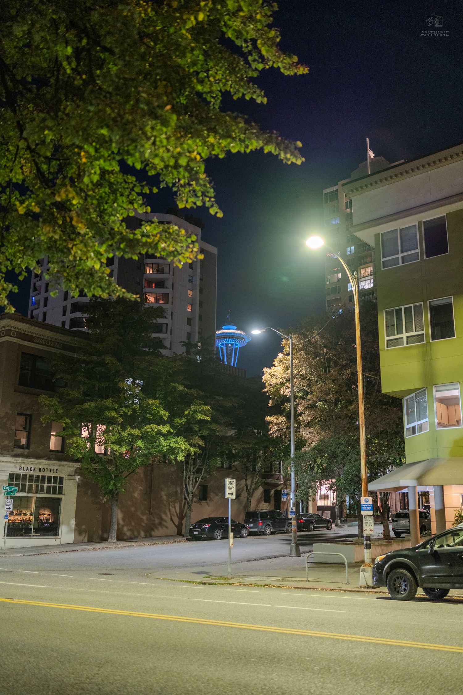 Night street scene with glowing Space Needle, lit streetlamps, green trees, and urban buildings in Seattle.