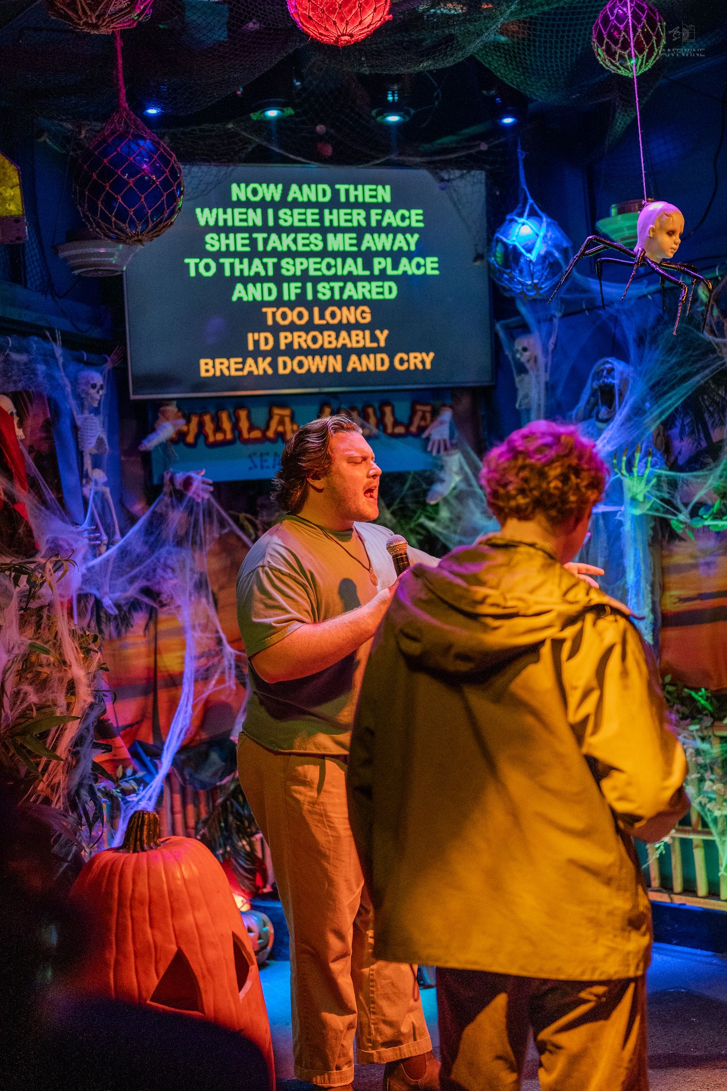 Two people sing karaoke at a Halloween-themed venue with spooky decorations and colorful lyrics displayed on screen.