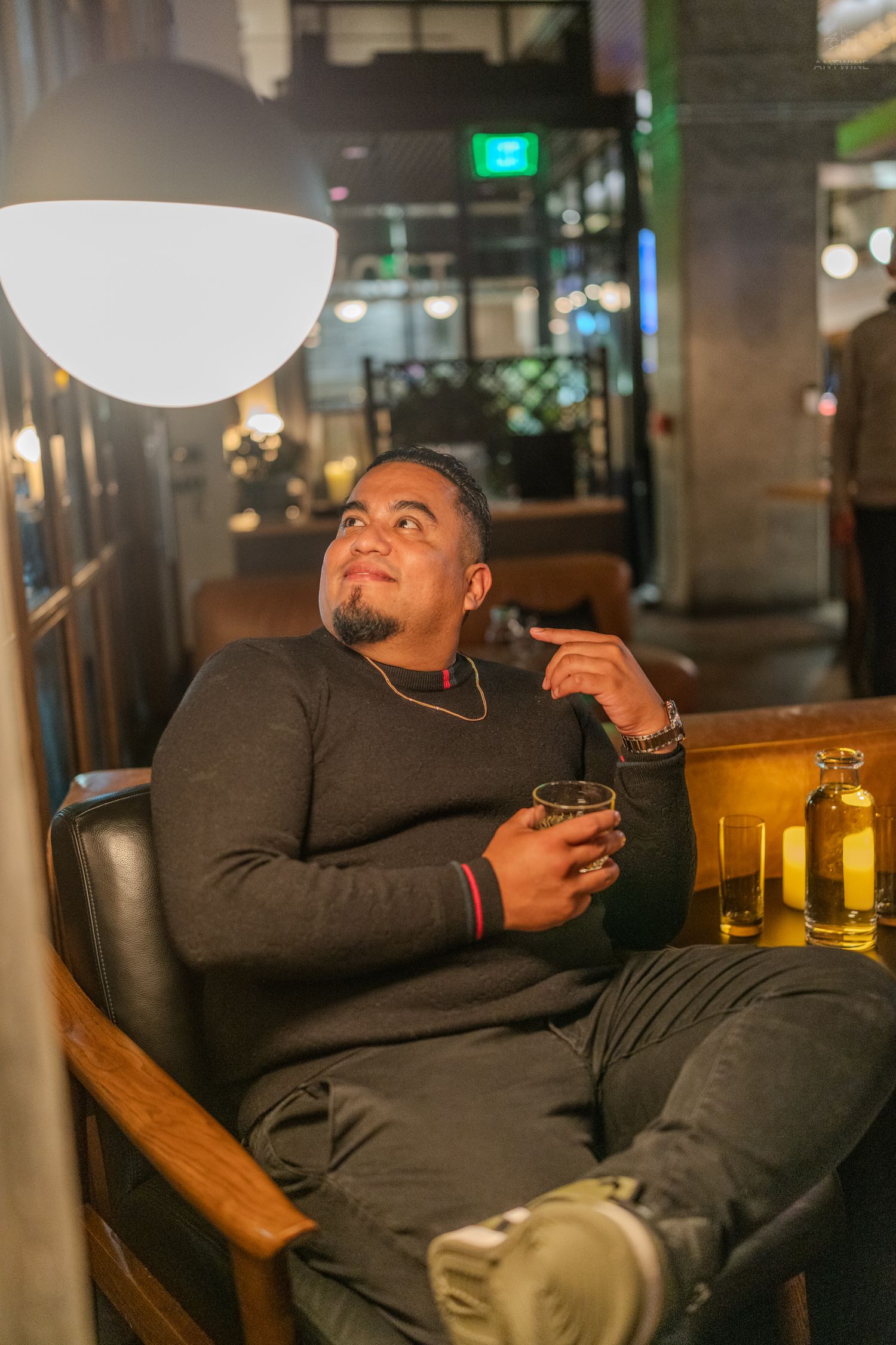 Man in black sweater relaxing in leather chair at bar, holding whiskey glass, looking up at pendant light.