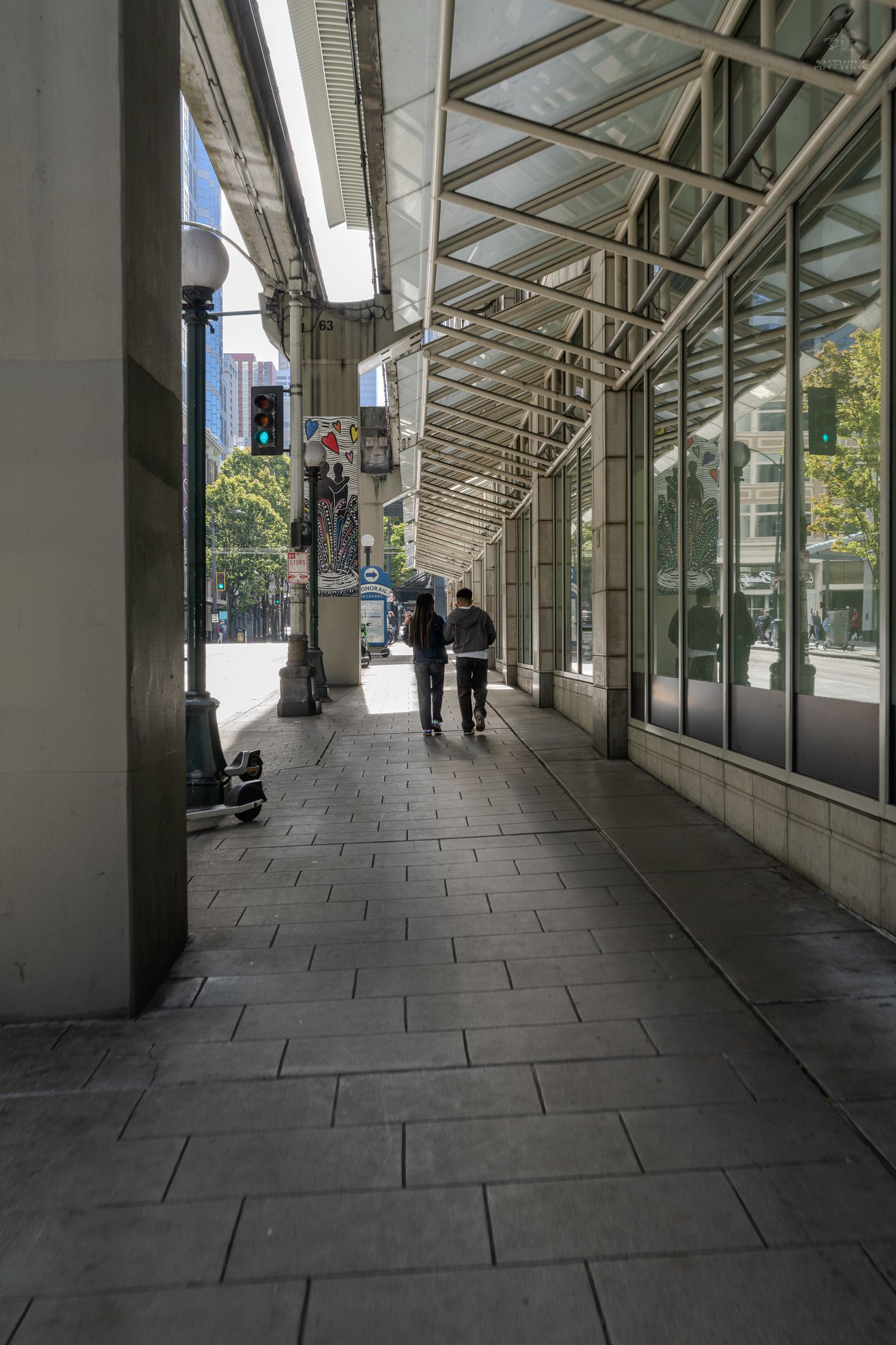 Two people walking along a covered urban sidewalk with glass canopy and stone pavement in a city.