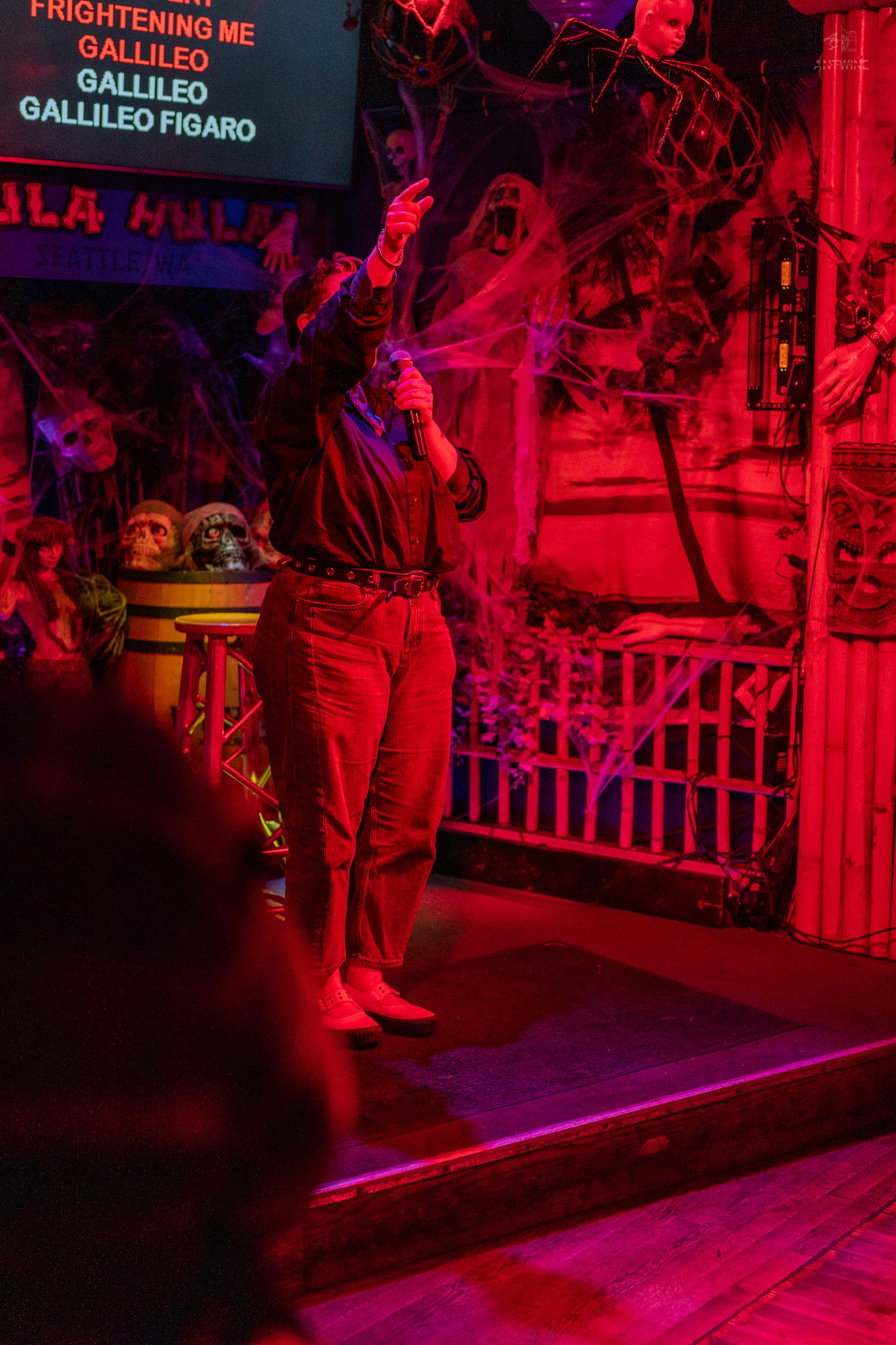 Person singing Bohemian Rhapsody karaoke on a Halloween-decorated stage bathed in red light.