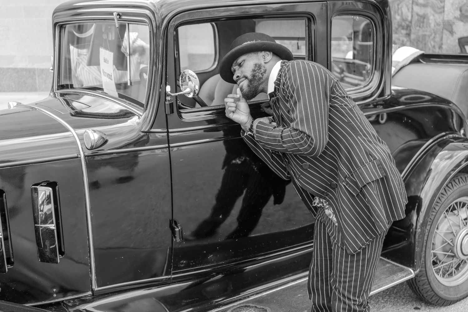 A man in a pinstripe suit examines a vintage black classic car in a black and white photograph.