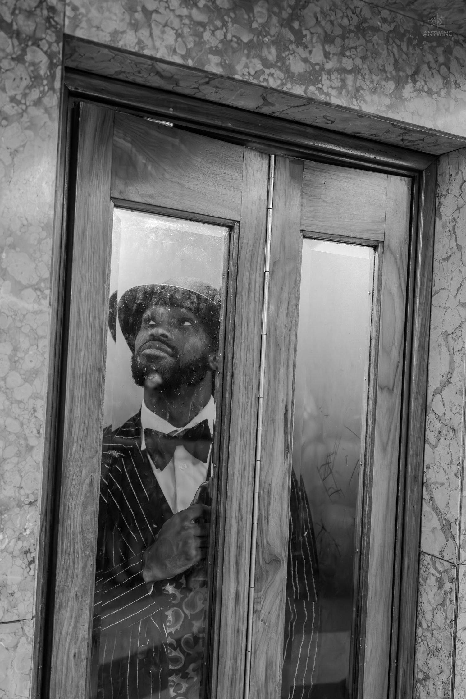 Black and white photo of a man in a pinstripe suit and bow tie peering through a glass door.