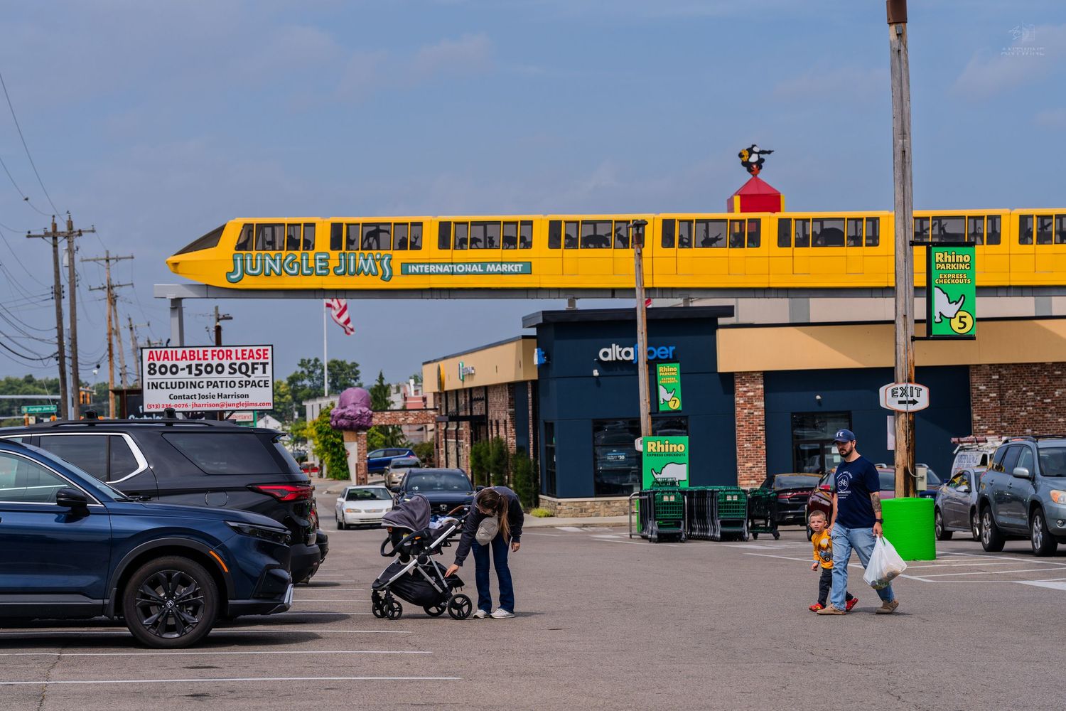 Yellow monorail train passes over a busy strip mall parking lot with shoppers and strollers on a sunny day.