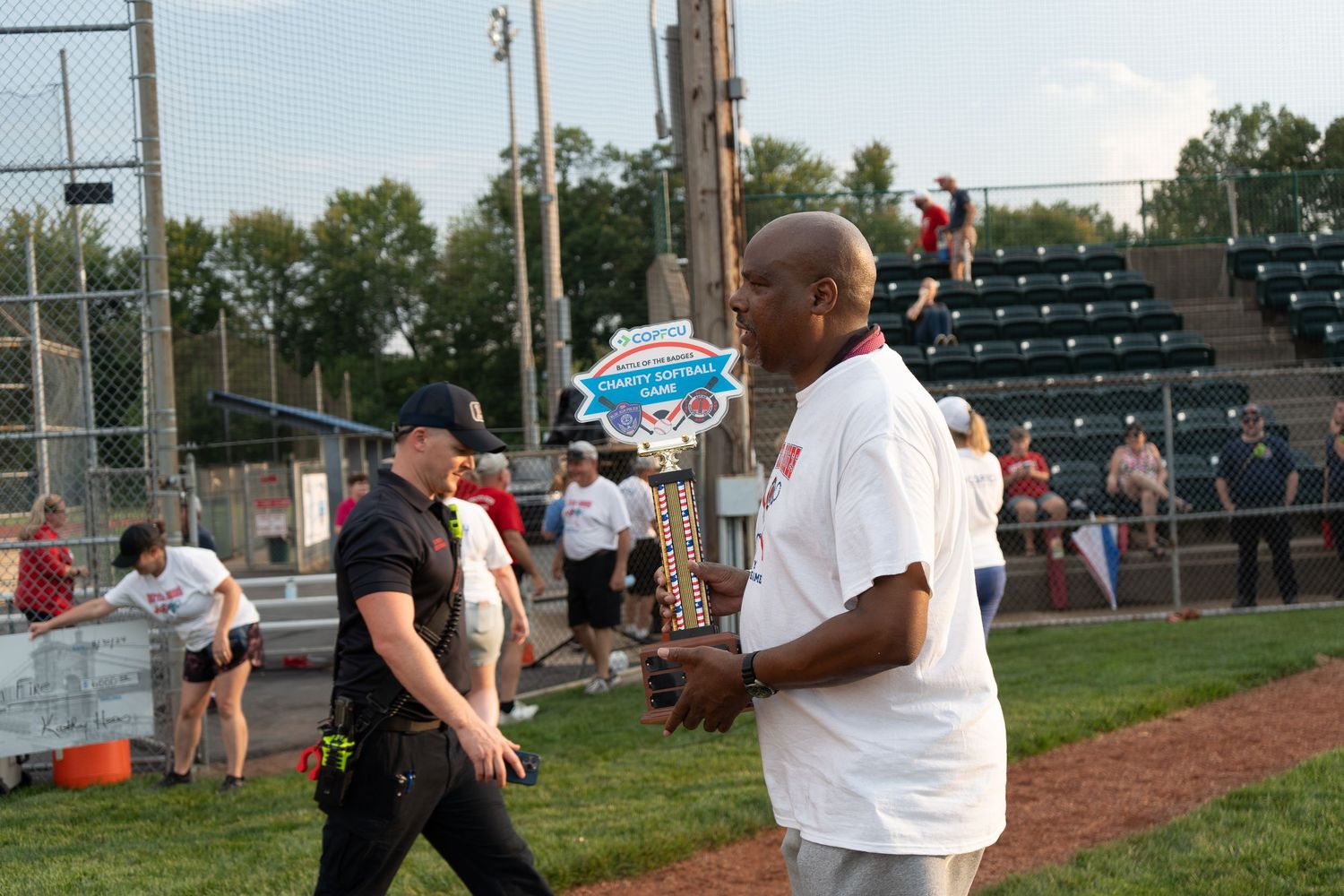 Man holds championship trophy at baseball field with crowd in background.