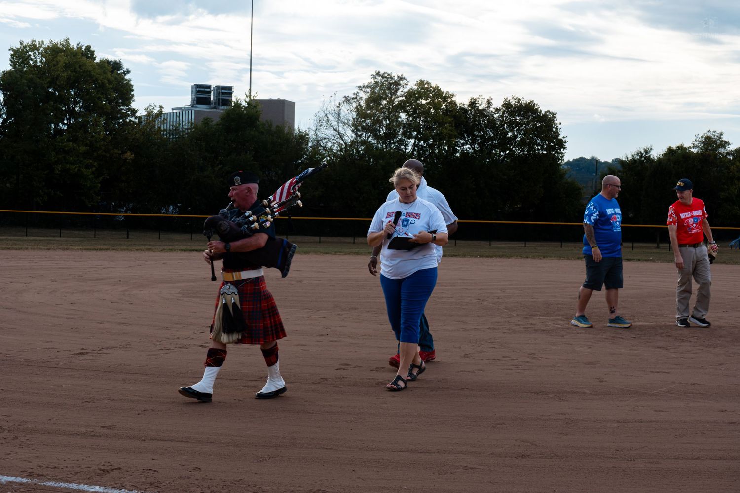 Bagpiper in tartan kilt leads a group across a dirt baseball field carrying an American flag at dusk.