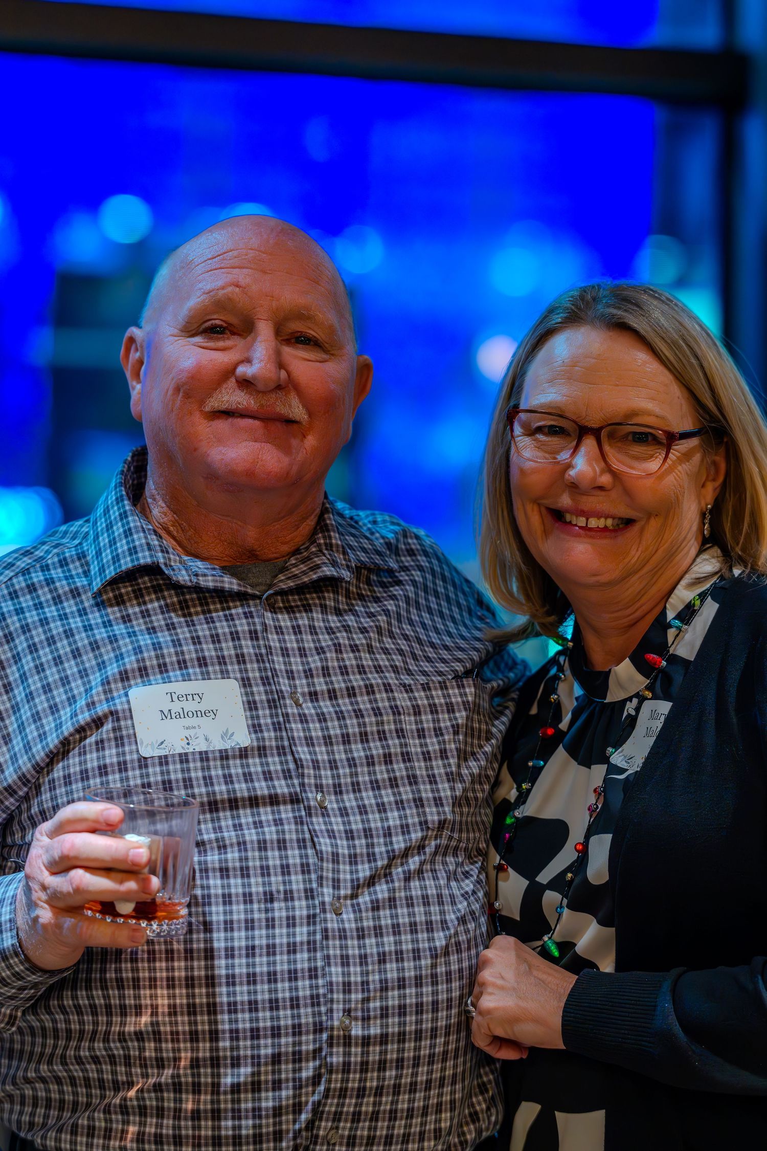 Two smiling adults pose together at a social event, man holding a drink wearing a name tag, woman in patterned top with glasses.