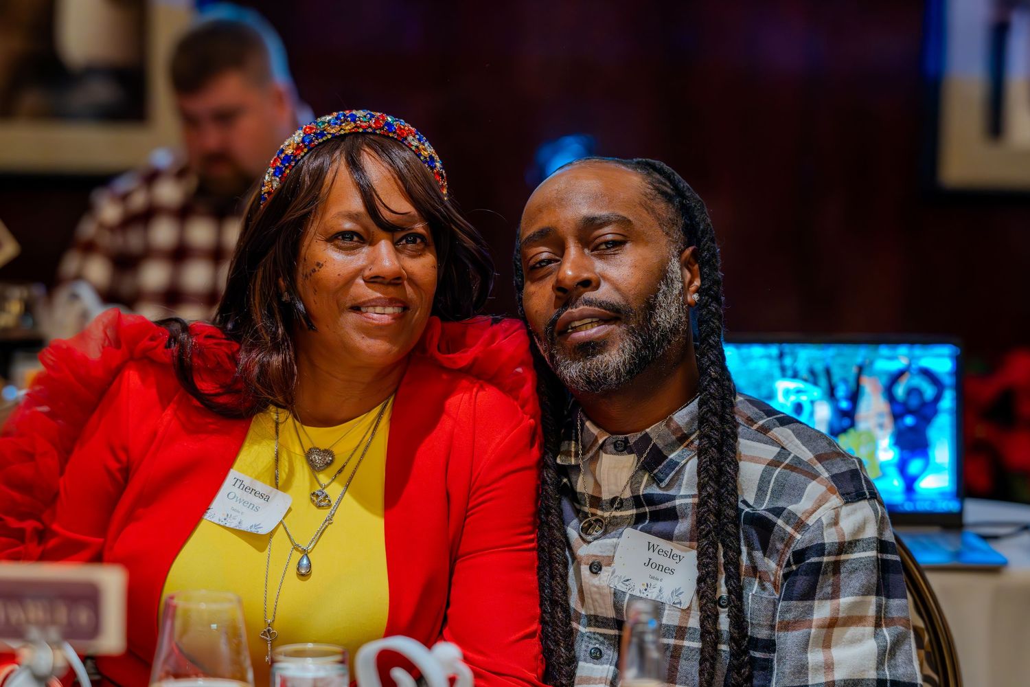 Two people smiling and posing together at an indoor event, woman in red jacket and man with dreadlocks.