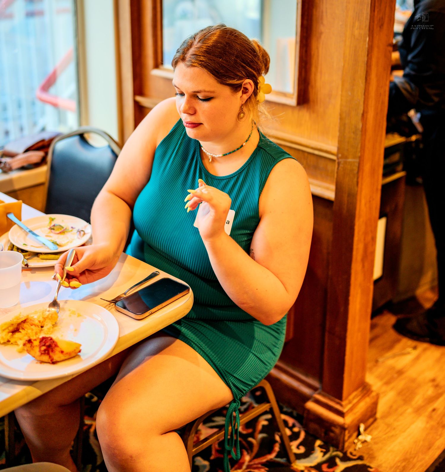 Woman in teal dress dining at restaurant table with food plate and smartphone nearby.