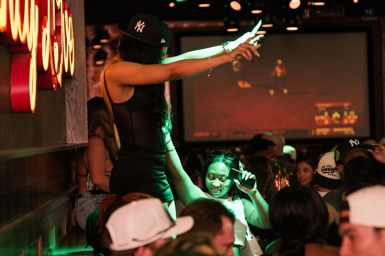 Woman dances on bar as crowd celebrates under green lights at lively sports bar.
