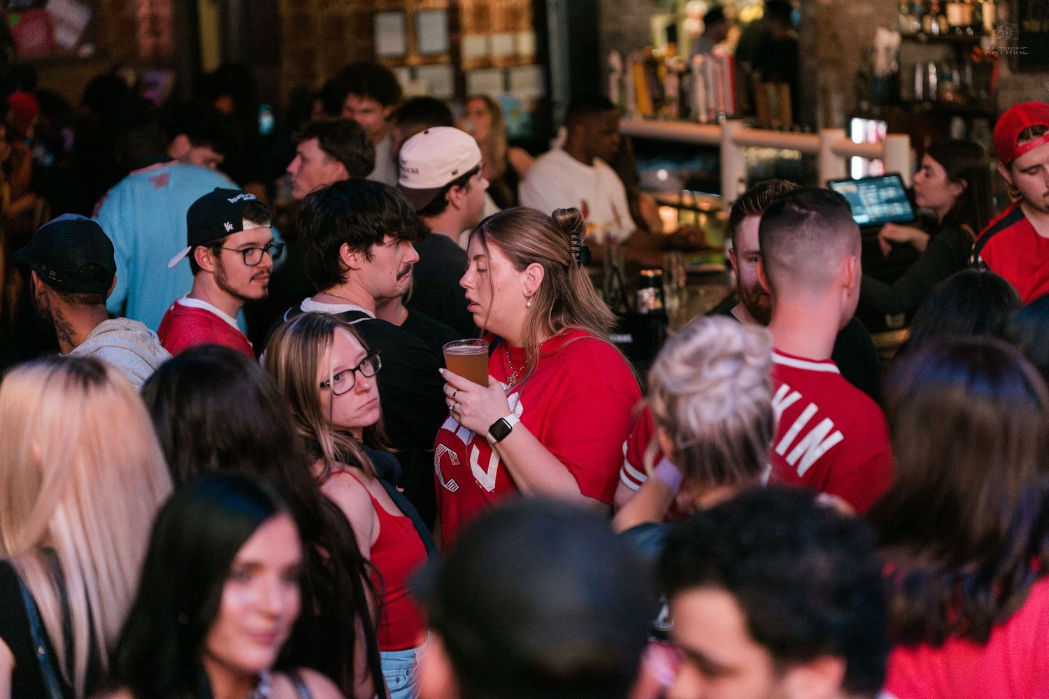 A crowd of people socialize and hold drinks at a busy, red-lit bar or nightclub event.