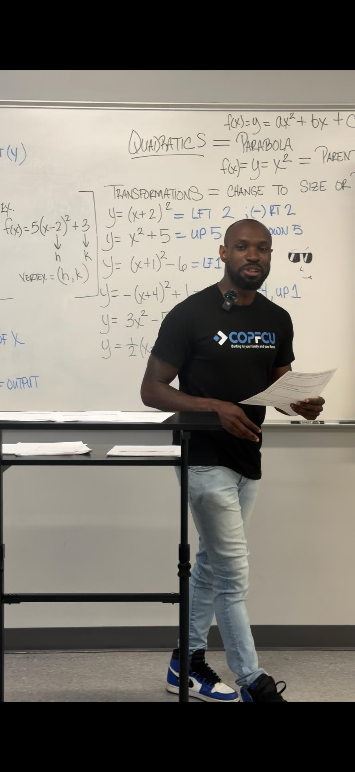 A math teacher in a COPFCU shirt stands at a podium presenting a quadratics lesson on a whiteboard in a classroom.