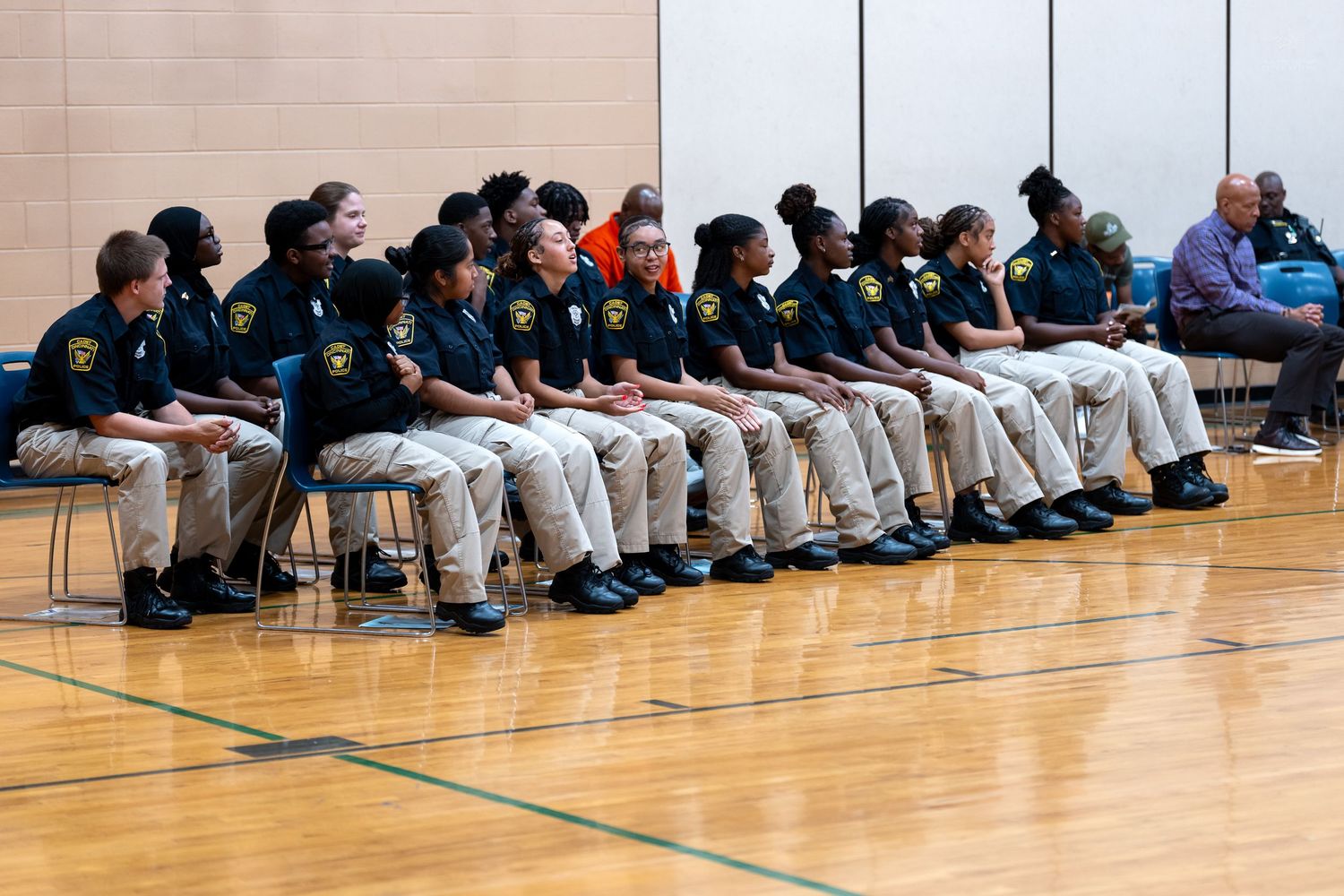 Youth officers in uniform shirts and khaki pants seated in rows on a gymnasium floor during a formal ceremony.
