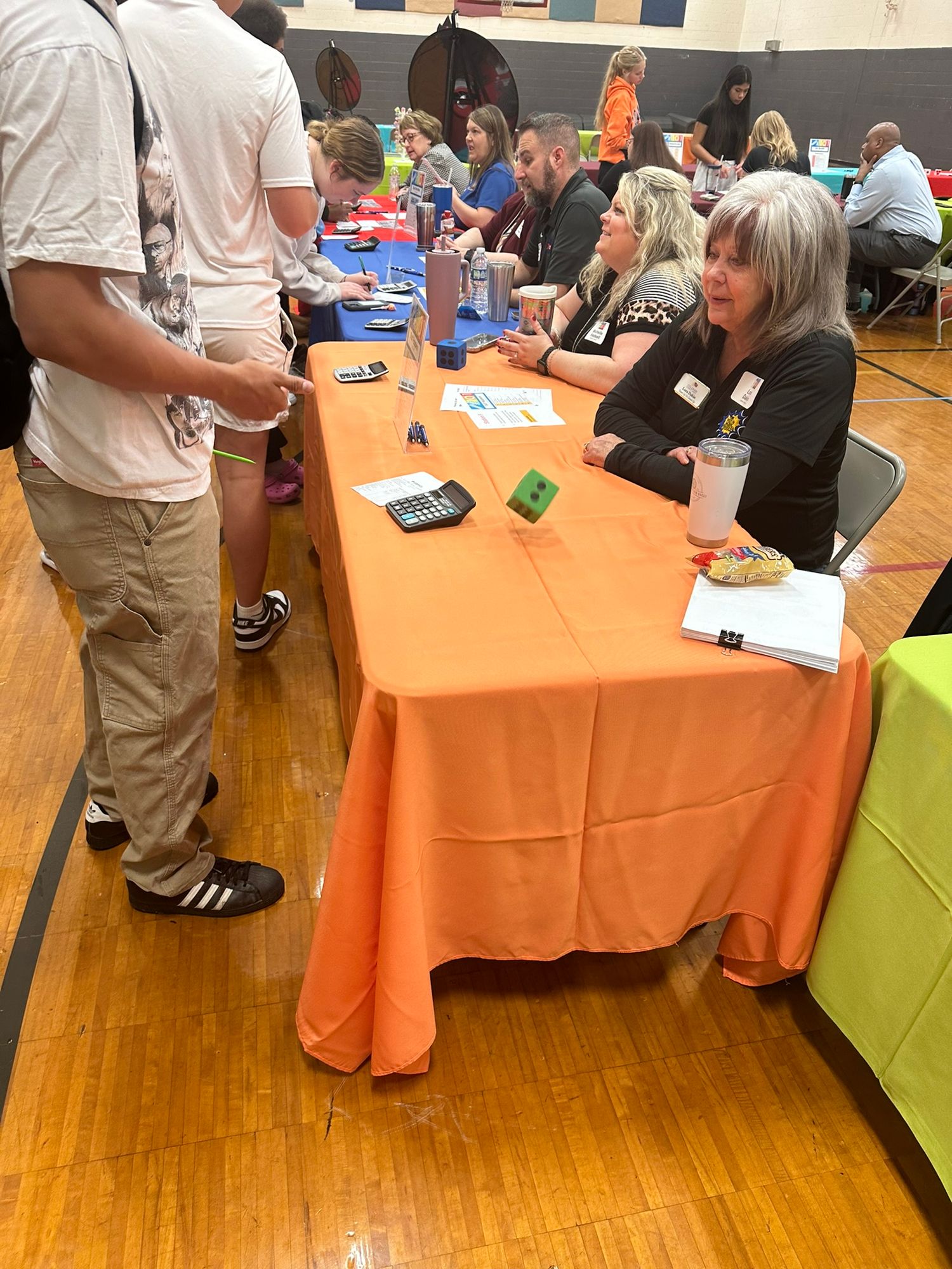 People networking at an indoor community event with orange tablecloths in a gymnasium setting.