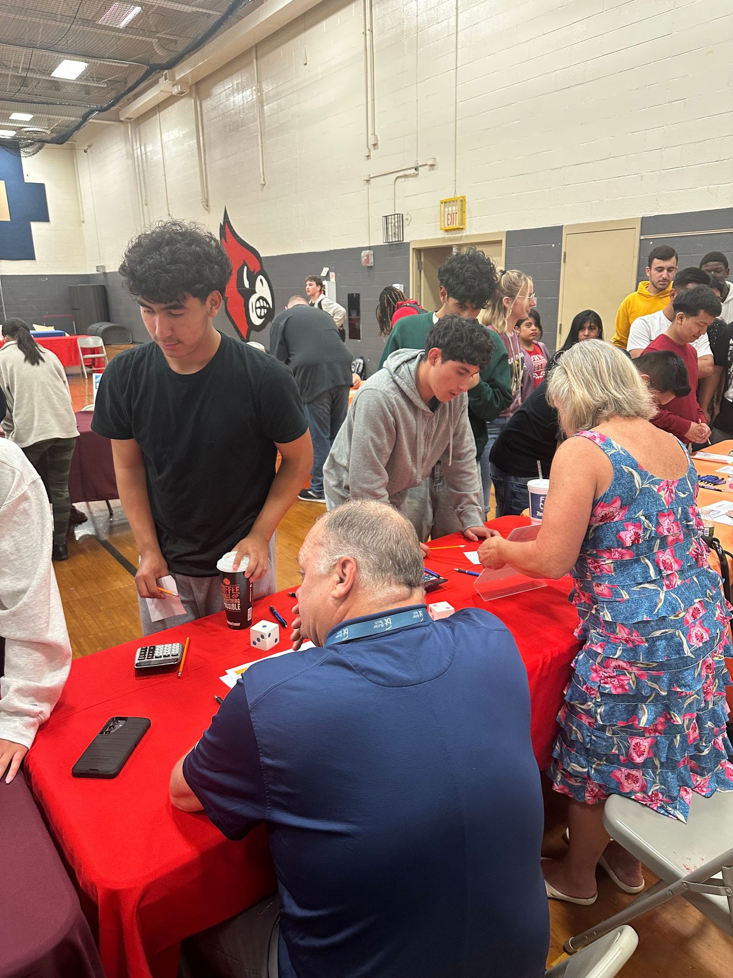 Students and adults gather around red tables in a school gymnasium during an organized event or fair.
