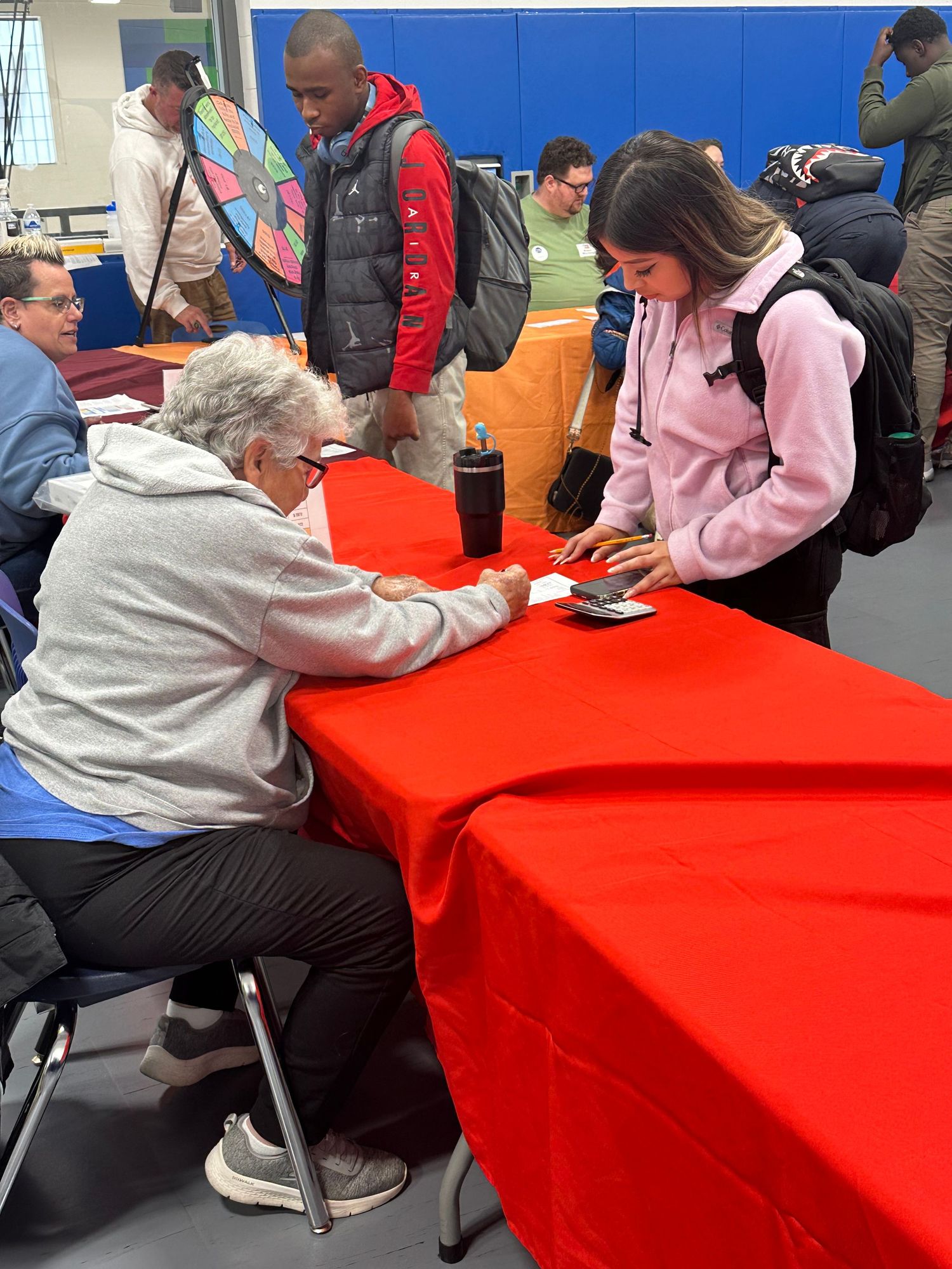 People gathered at a community event with red tablecloths, a woman in pink hoodie interacts with an elderly person at a table.
