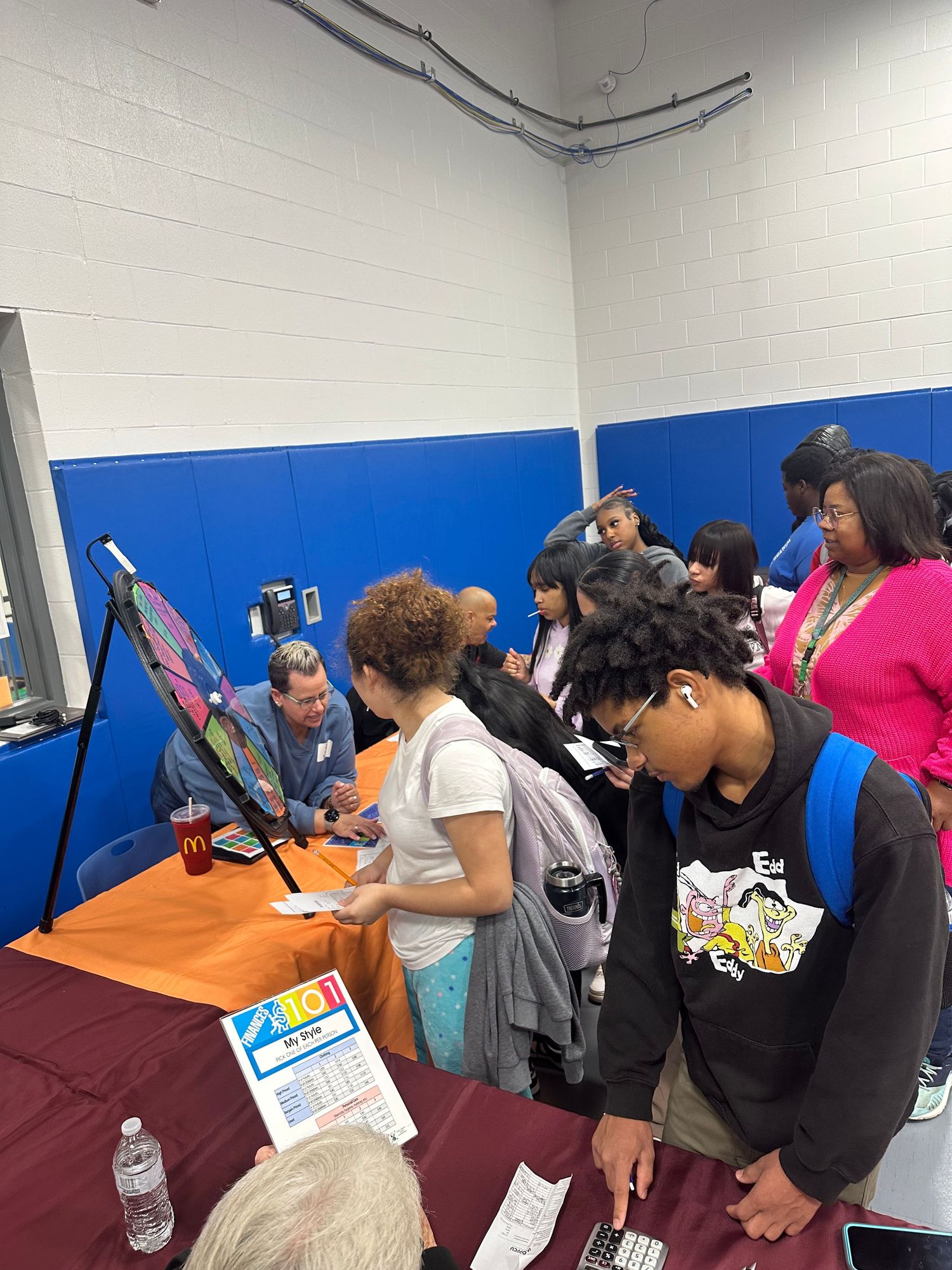 Students gather around orange and maroon tables interacting with representatives at a busy school gymnasium career fair.