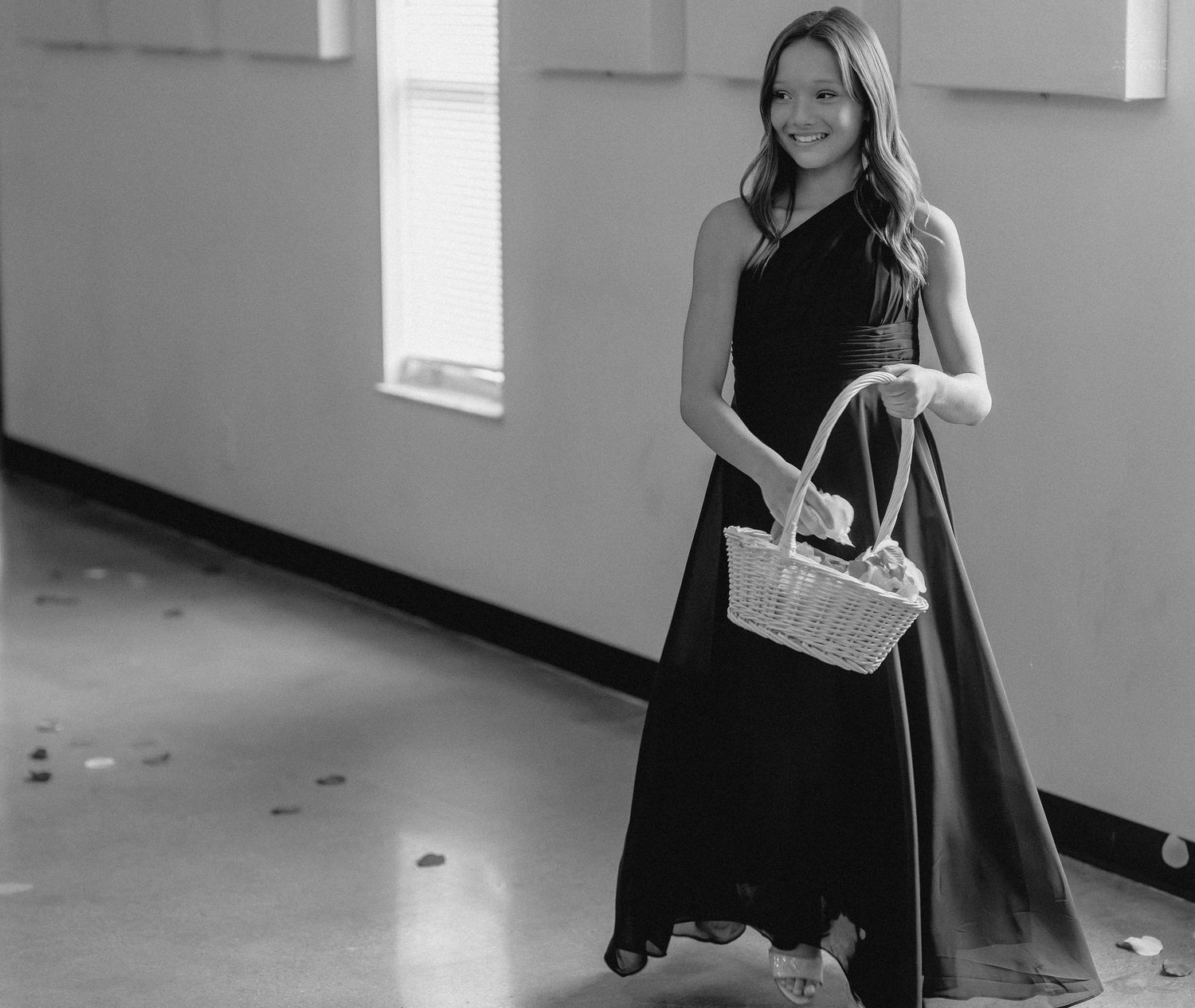Young woman in black gown holding wicker basket, smiling in black and white wedding photo.