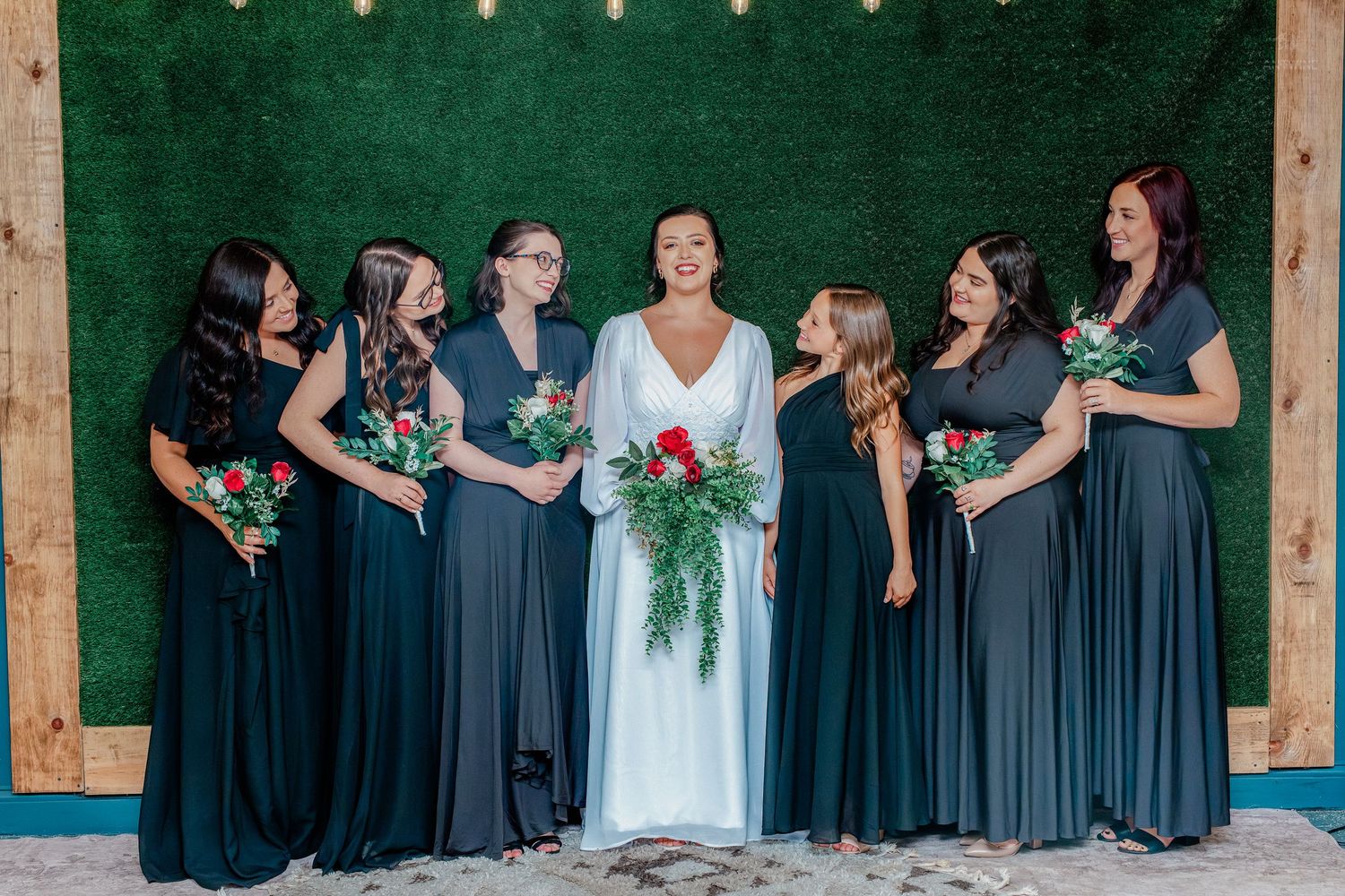 Bride in white gown poses with six bridesmaids in dark gray dresses holding red bouquets before a green hedge wall.