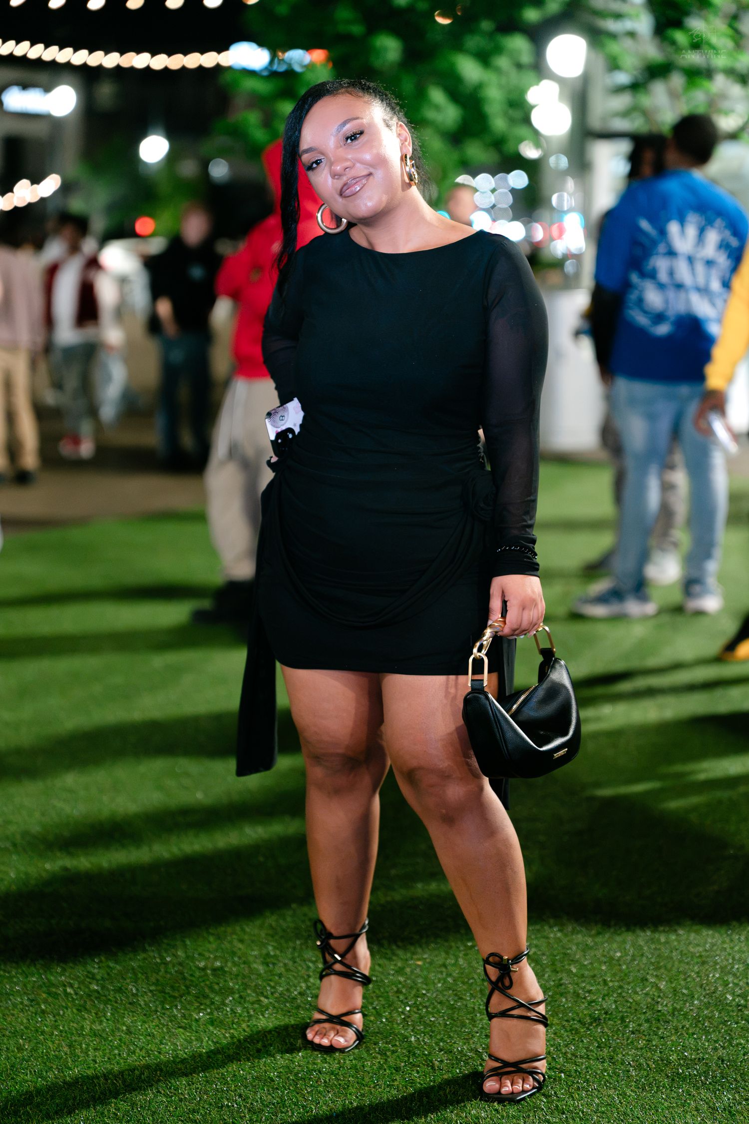 Smiling woman in black long sleeve mini dress holding purse at outdoor evening event.