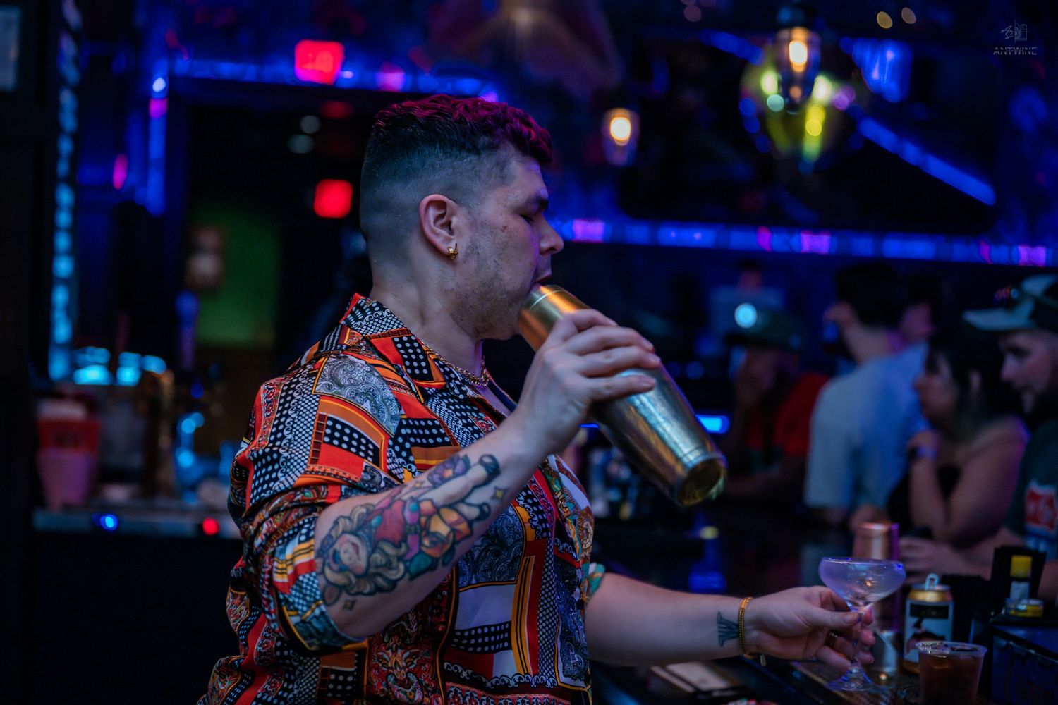 Tattooed man in patterned shirt drinks from shaker at a vibrant nightclub bar.